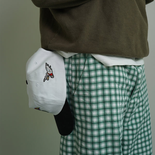Person wearing a white cap with a design, brown sweatshirt, and green plaid pants on a plain background
