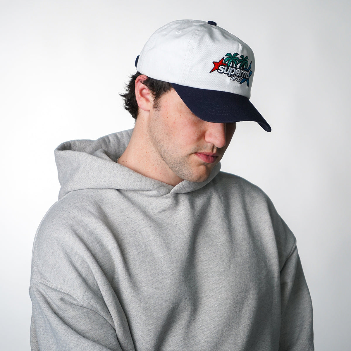 Person wearing a gray hoodie and a white and navy cap with a logo on a light gray background