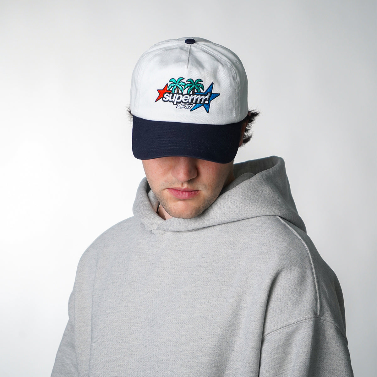 Person wearing a white cap with a colorful logo and a gray hoodie on a light gray background