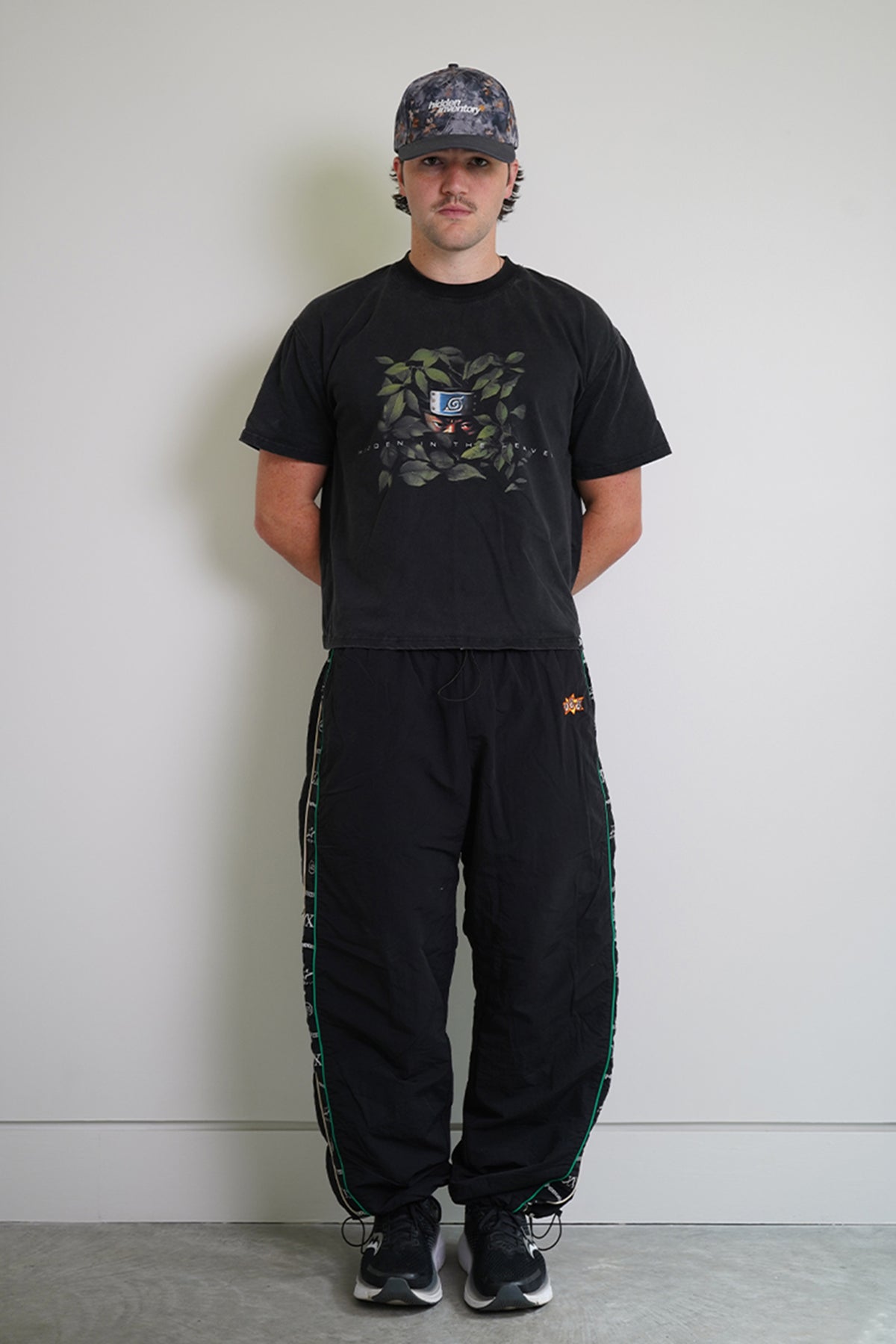Front view on model of the black Jan-ken Trackpants, baggy nylon with green piping, printed side stripes, drawstring ankle cuffs and a small orange embroidered logo
