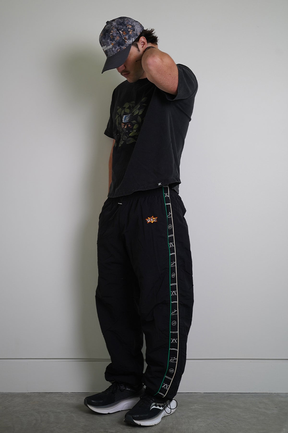 Model wearing the black Jan-ken Trackpants with green and cream side stripe of repeating woven logos, small orange embroidered emblem, side view