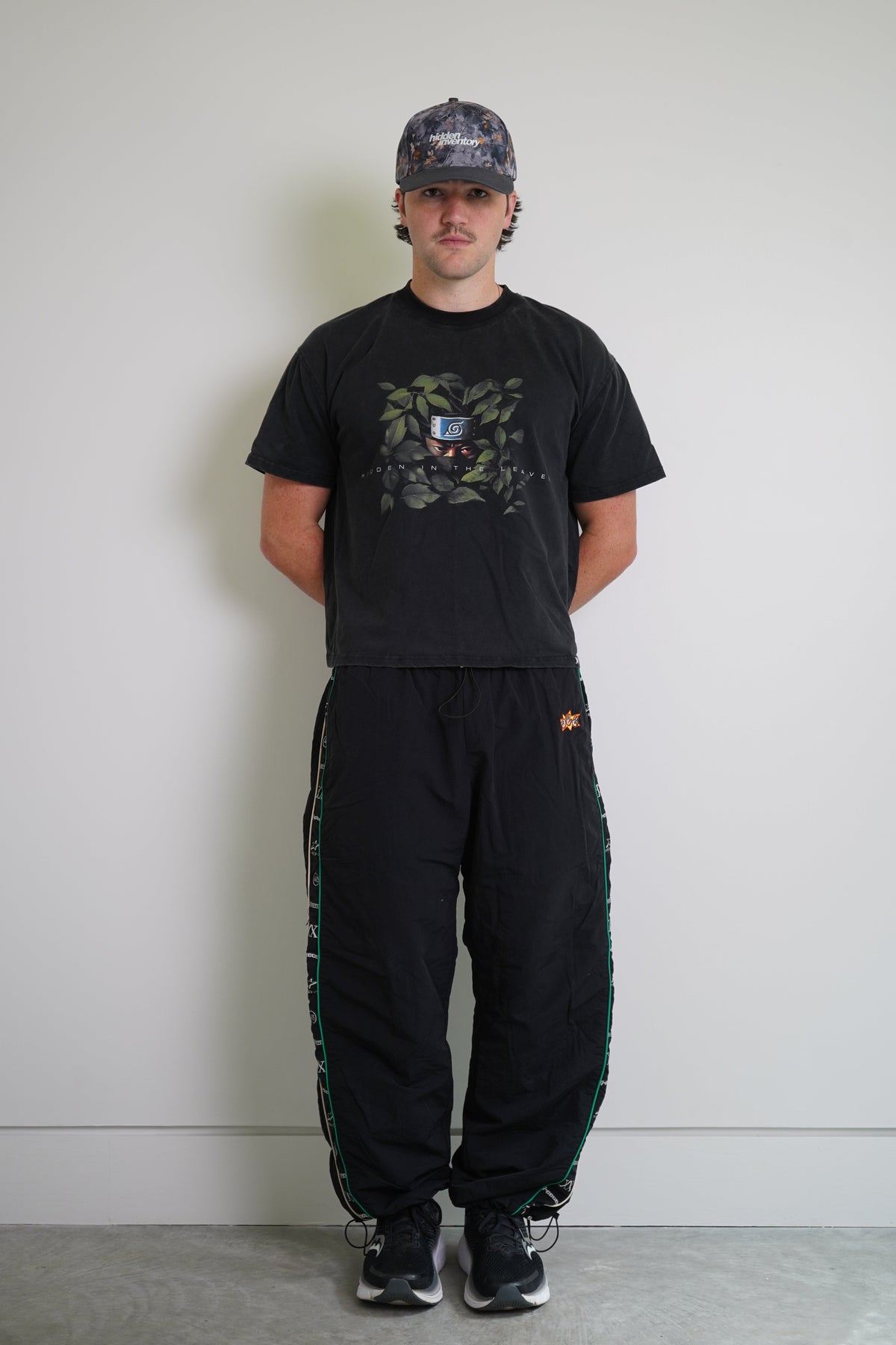 A man stands against a plain light gray wall wearing a black graphic t-shirt, black sweatpants with green side stripes, and a dark camouflage baseball cap.