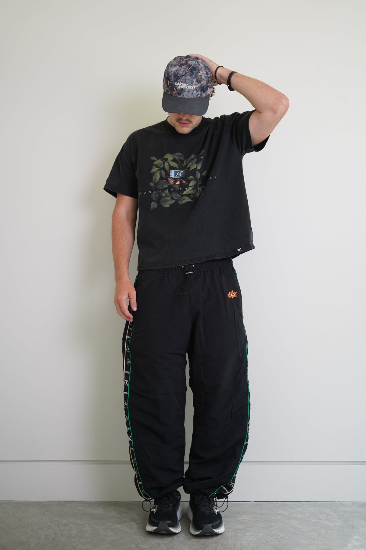 A person modeling a black graphic t-shirt, black sweatpants with green side stripes, and a camouflage baseball cap against a plain white wall.