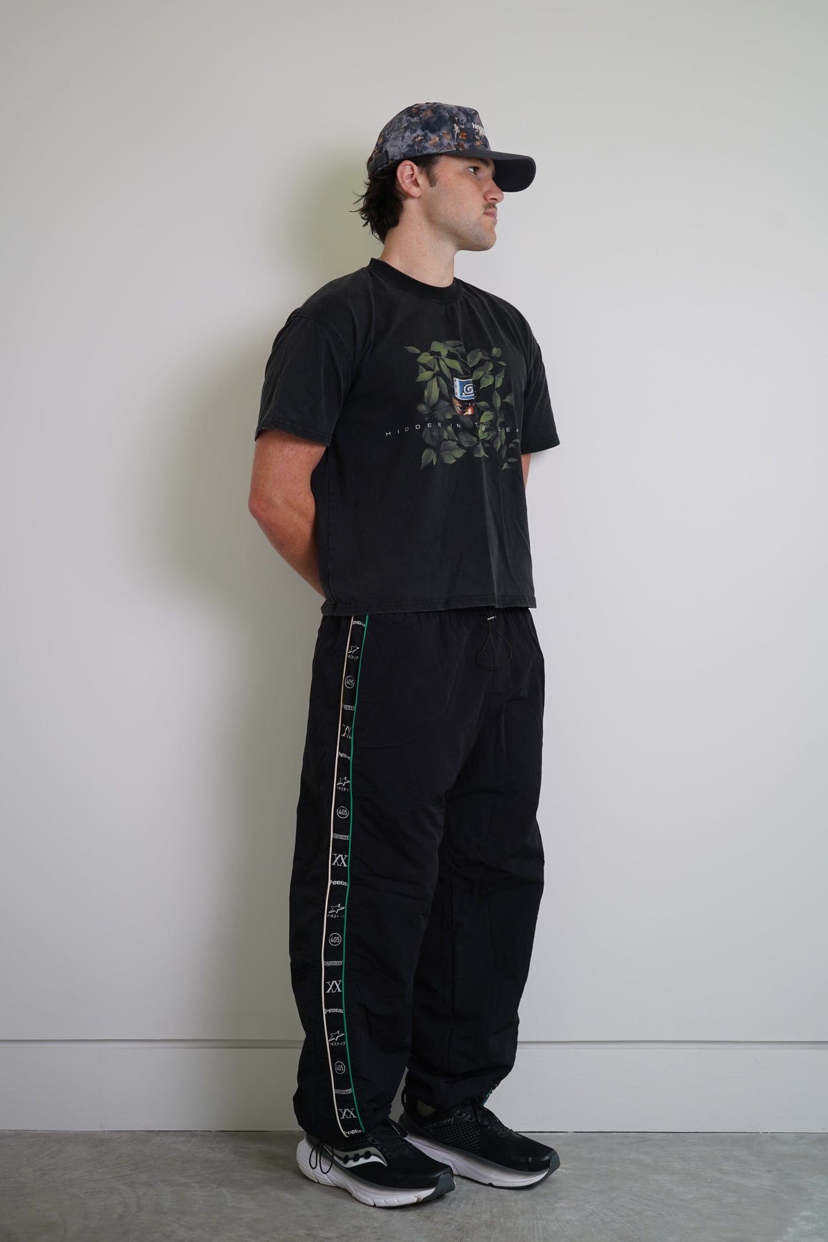 A man stands in profile wearing a black graphic t-shirt, black sweatpants with a green and white side stripe, a dark patterned baseball cap, and black sneakers, set against a plain light gray wall.