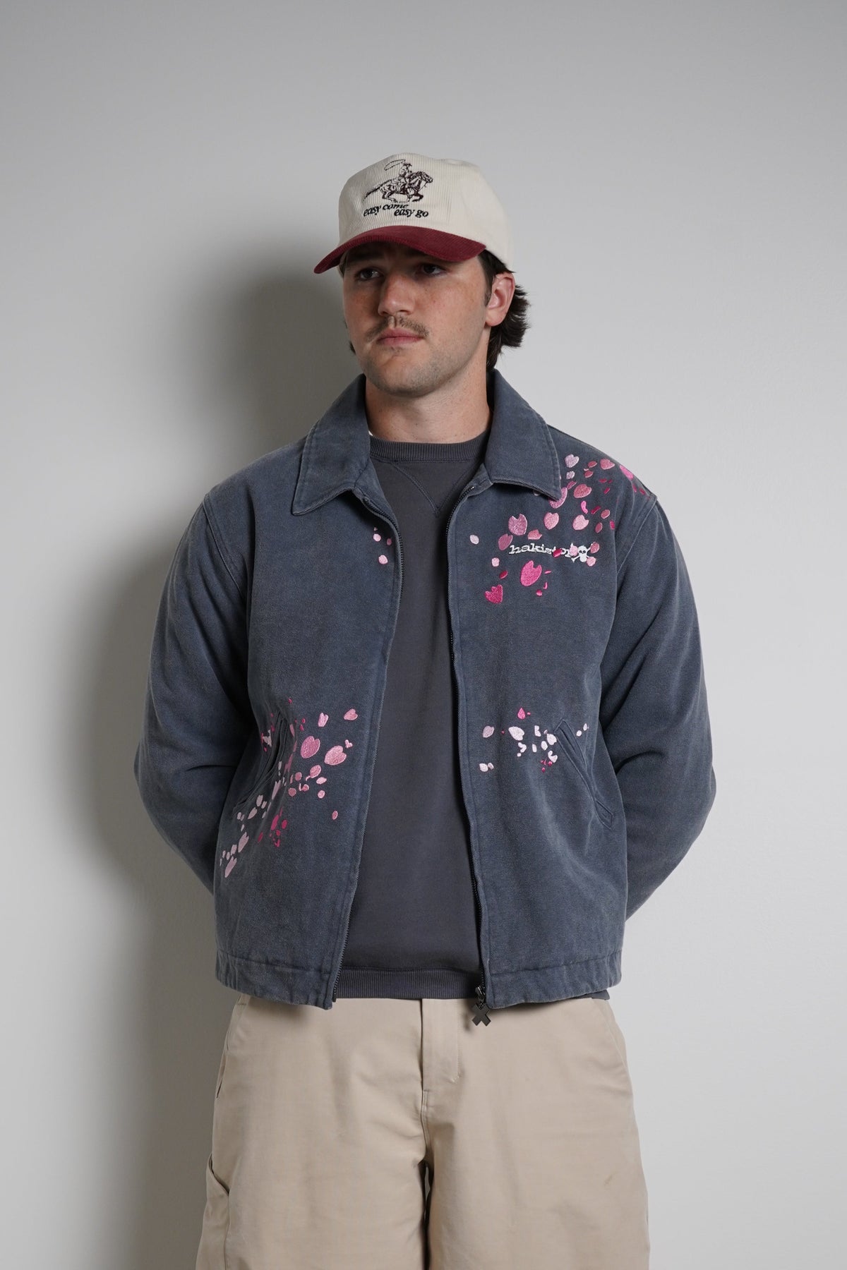 A man stands against a plain light gray wall wearing a faded blue cropped jacket with pink and white floral embroidery, beige cargo pants, a dark gray t-shirt, and a beige baseball cap.