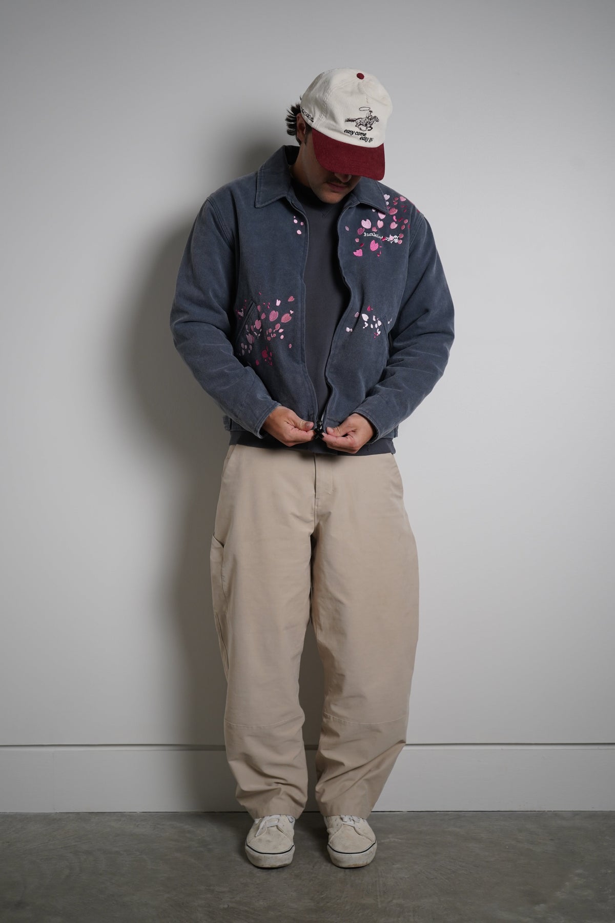 A person stands against a plain light gray wall wearing a dark blue zip-up jacket with pink floral embroidery, beige cargo pants, a white baseball cap with a red brim, and white sneakers.