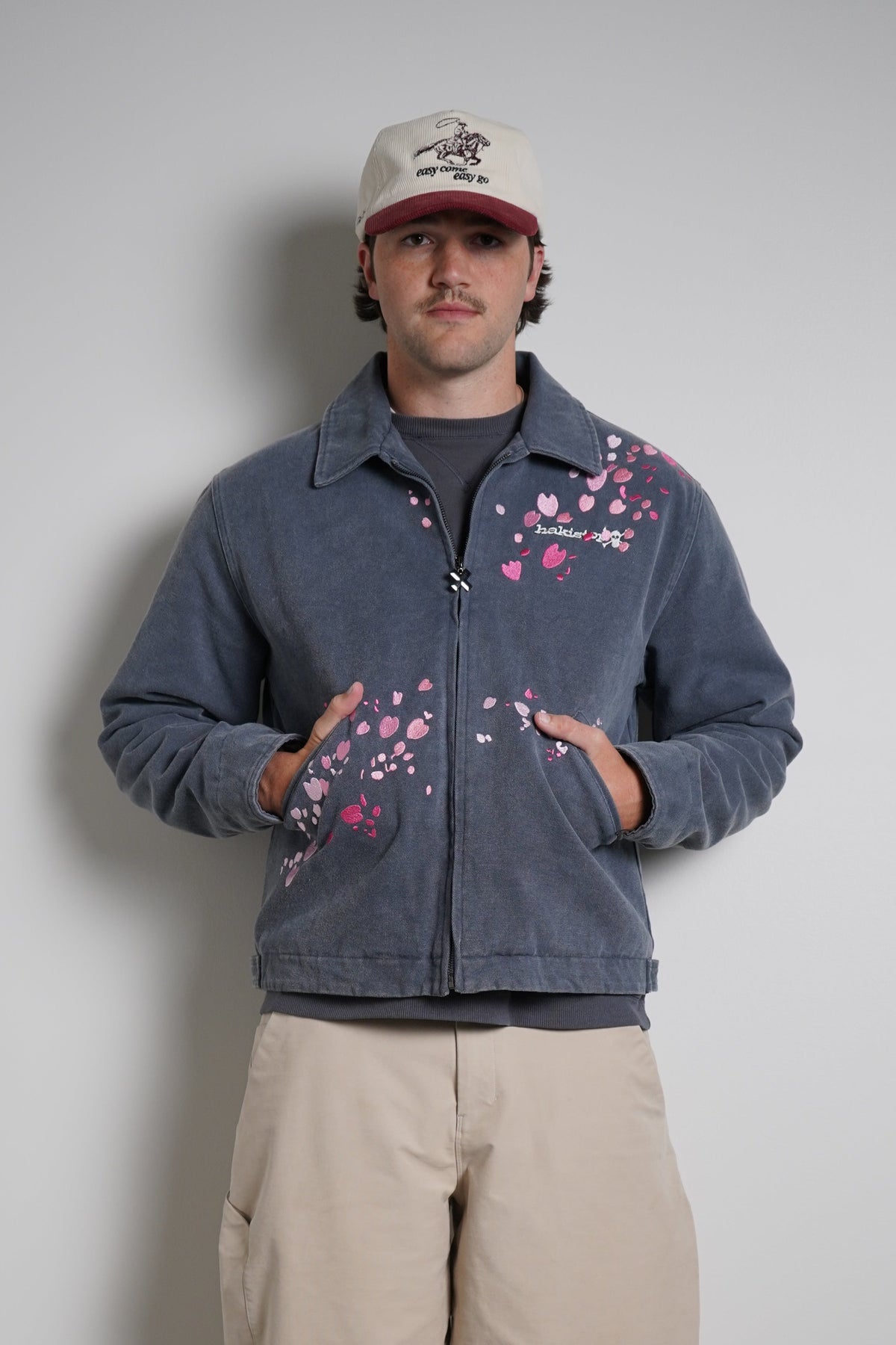 A man stands against a plain light gray wall wearing a dark blue cropped zip-up jacket with pink floral embroidery, beige cargo pants, white sneakers, and a beige baseball cap.