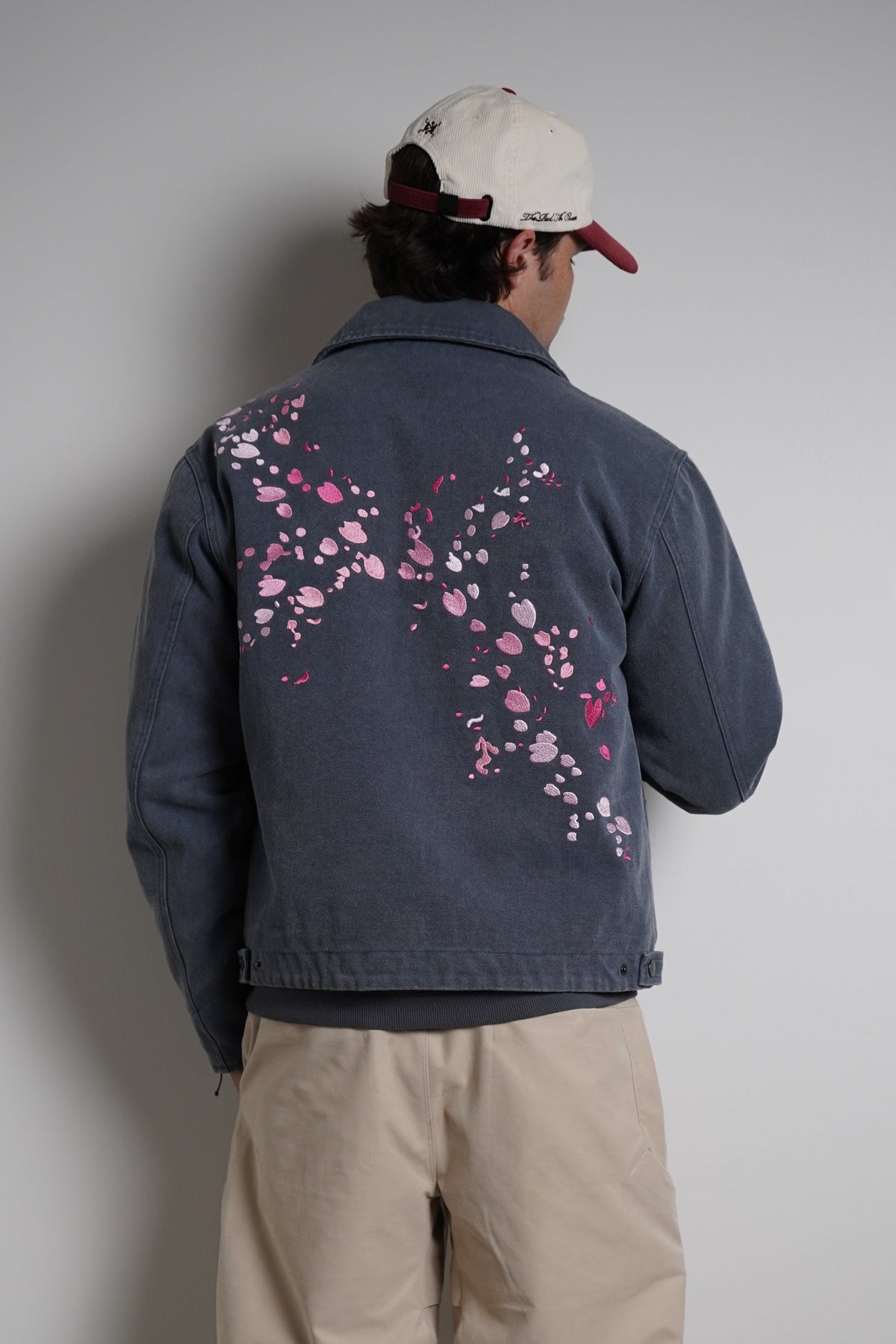 A person standing with their back to the camera, wearing a navy blue denim jacket with pink cherry blossom embroidery on the back, paired with beige pants and a white baseball cap.