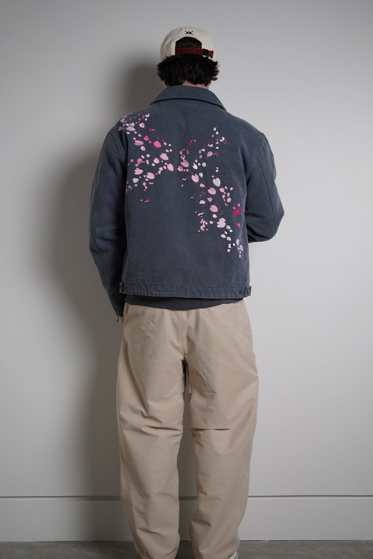 A person viewed from behind wearing a faded blue denim jacket with pink cherry blossom embroidery on the back, paired with beige pants and a beige cap, standing against a plain gray wall.