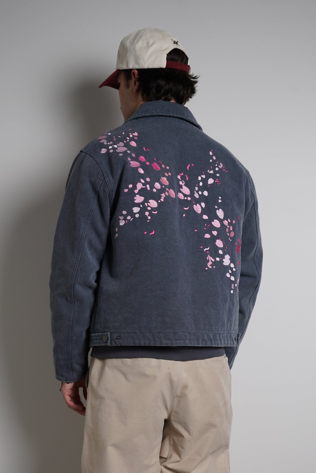 A person standing with their back to the camera, wearing a faded blue denim jacket with pink cherry blossom embroidery on the back, beige pants, and a cream baseball cap.