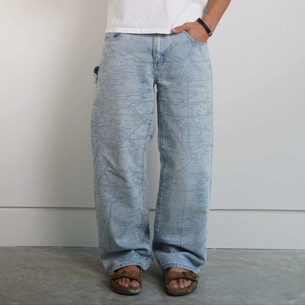A man wearing a white graphic t-shirt, wide-leg light blue jeans, and brown leather sandals, holding a phone to take a mirror selfie against a plain light gray wall.