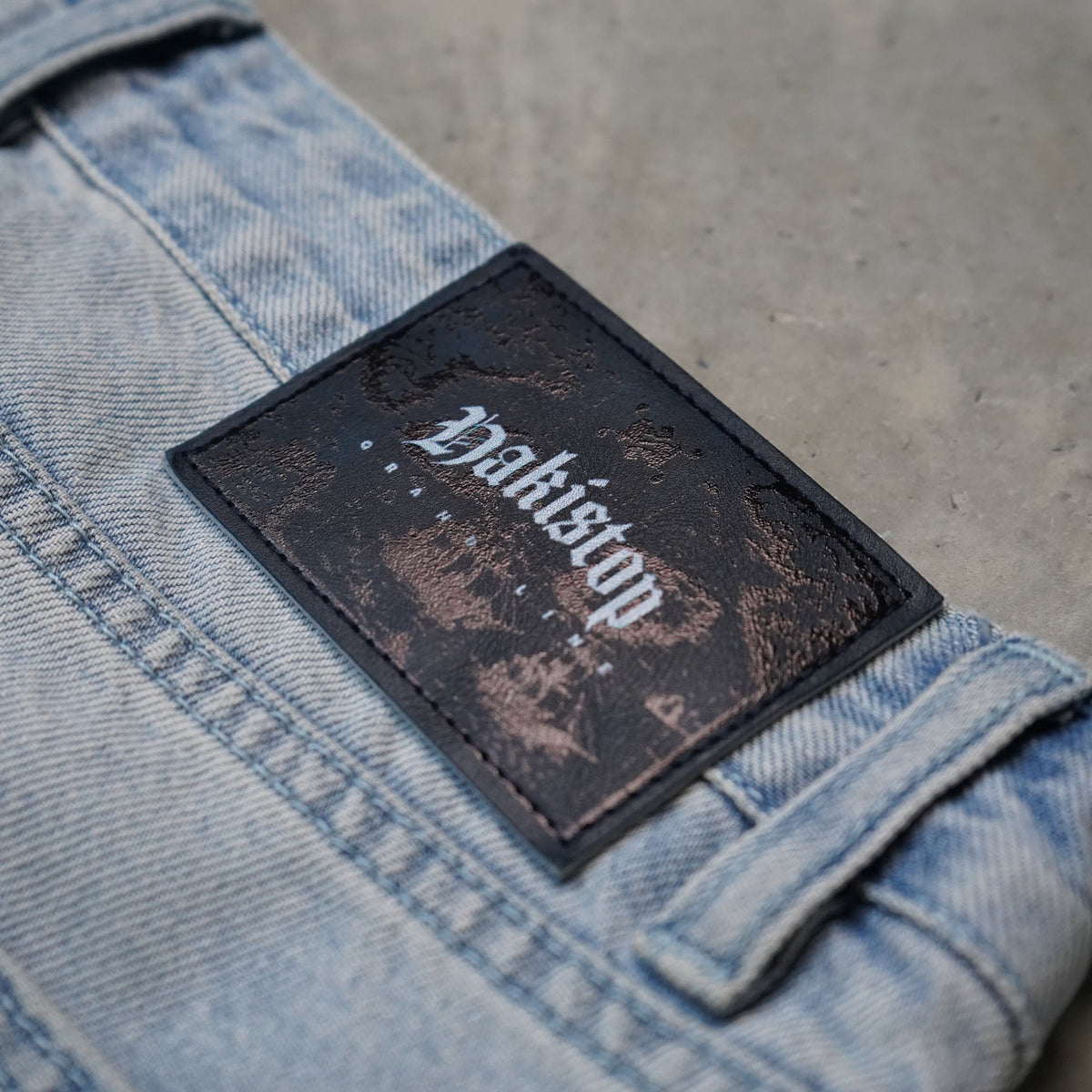 A close-up of light blue denim jeans showing a dark brown rectangular patch with the brand name 'Hakistop' in white gothic lettering, set against a textured gray concrete background.