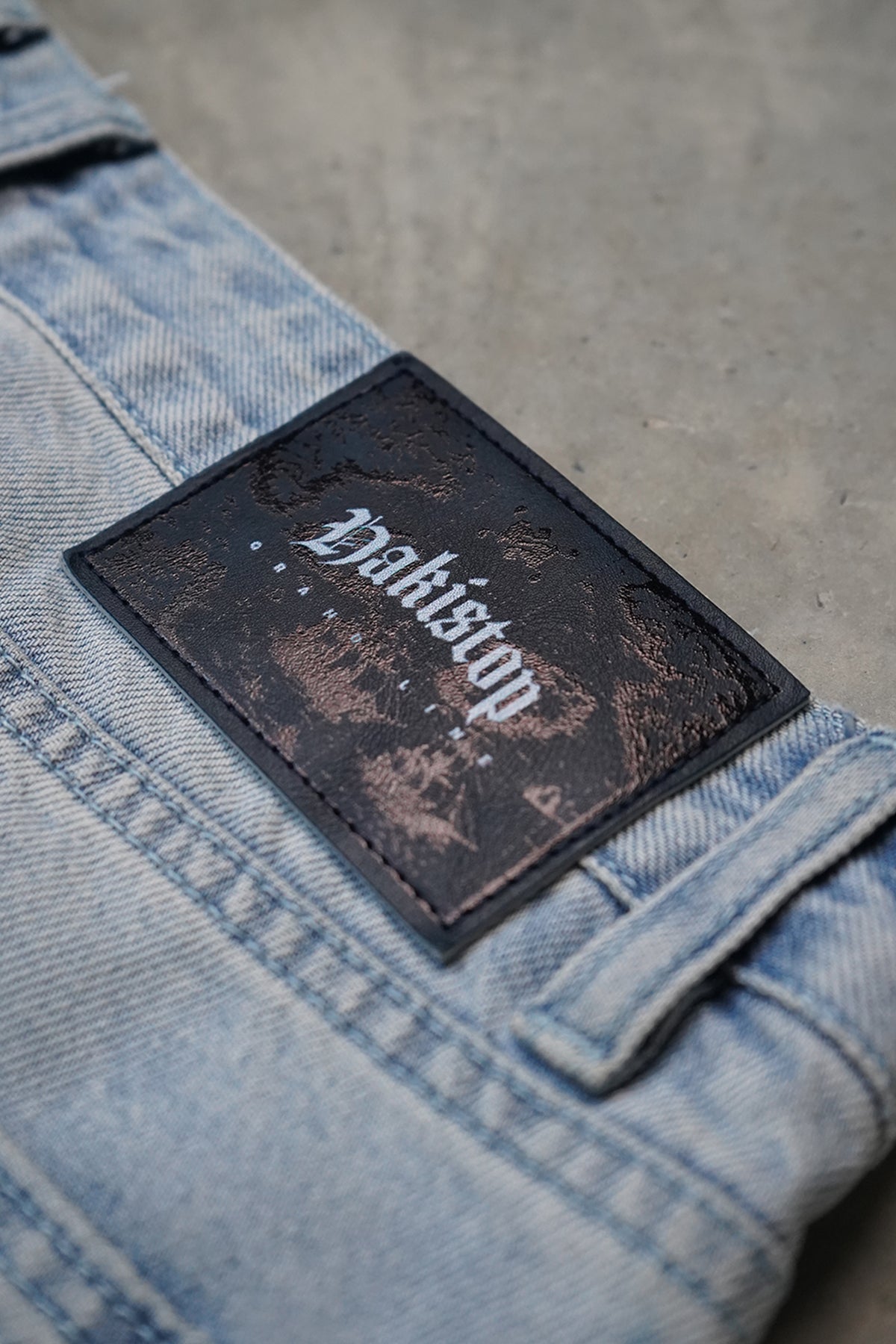 Close-up of the Grand Line Pants light blue washed denim with a black leather-look patch showing blackletter HAKISTOP text and a faded graphic