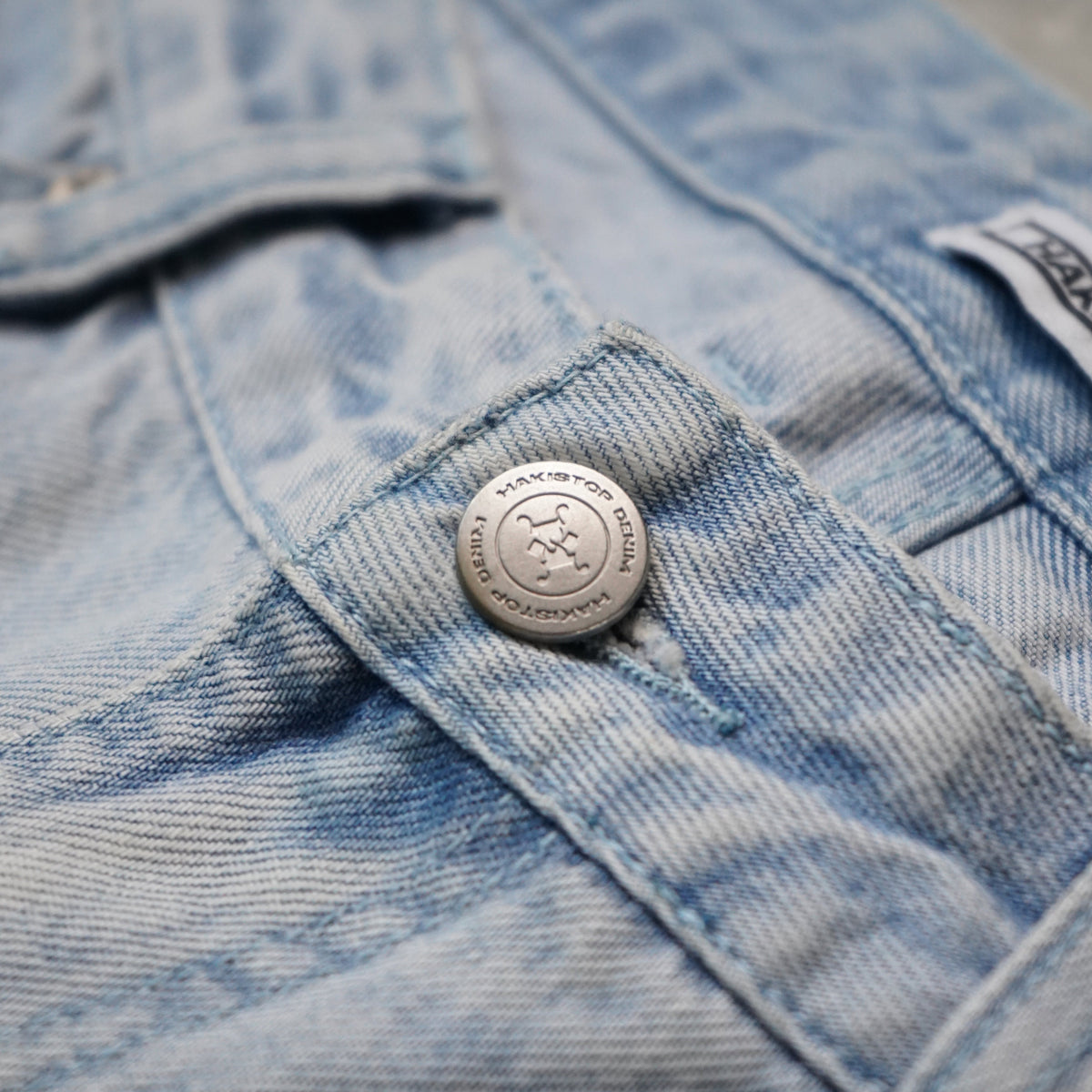 Close-up of a light blue denim garment, showing the waistband, button closure, and a small fabric brand tab on a gray background.