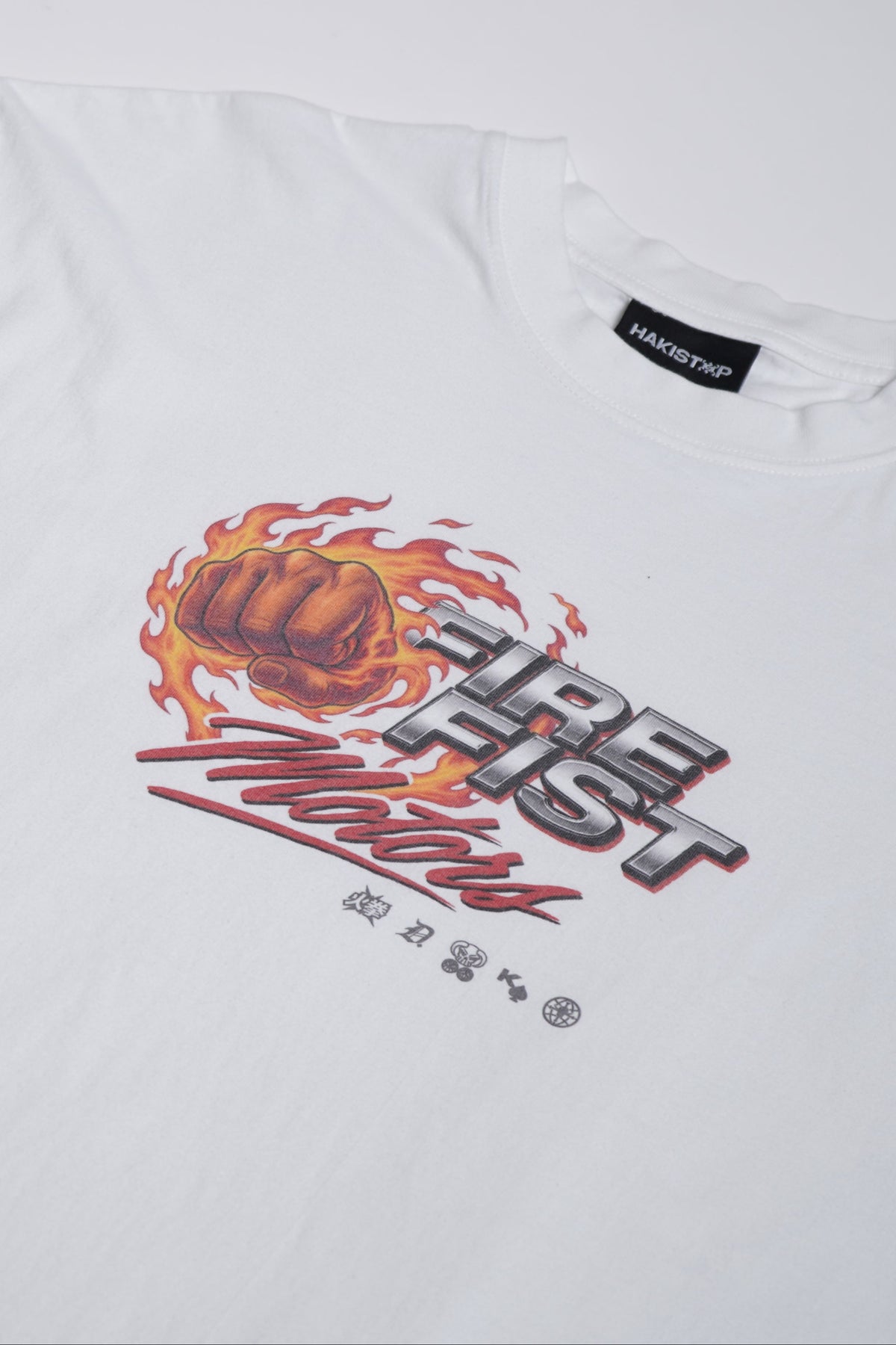 A white short-sleeve t-shirt laid flat on a light gray background, featuring a graphic print on the left chest with a basketball, flames, and stylized text reading 'Fire Fist Motors'.