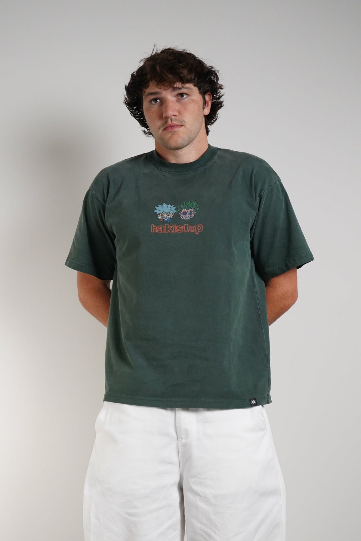A man wearing a dark green t-shirt with a small colorful graphic on the chest, paired with loose white pants and black sneakers, standing against a plain light gray wall.