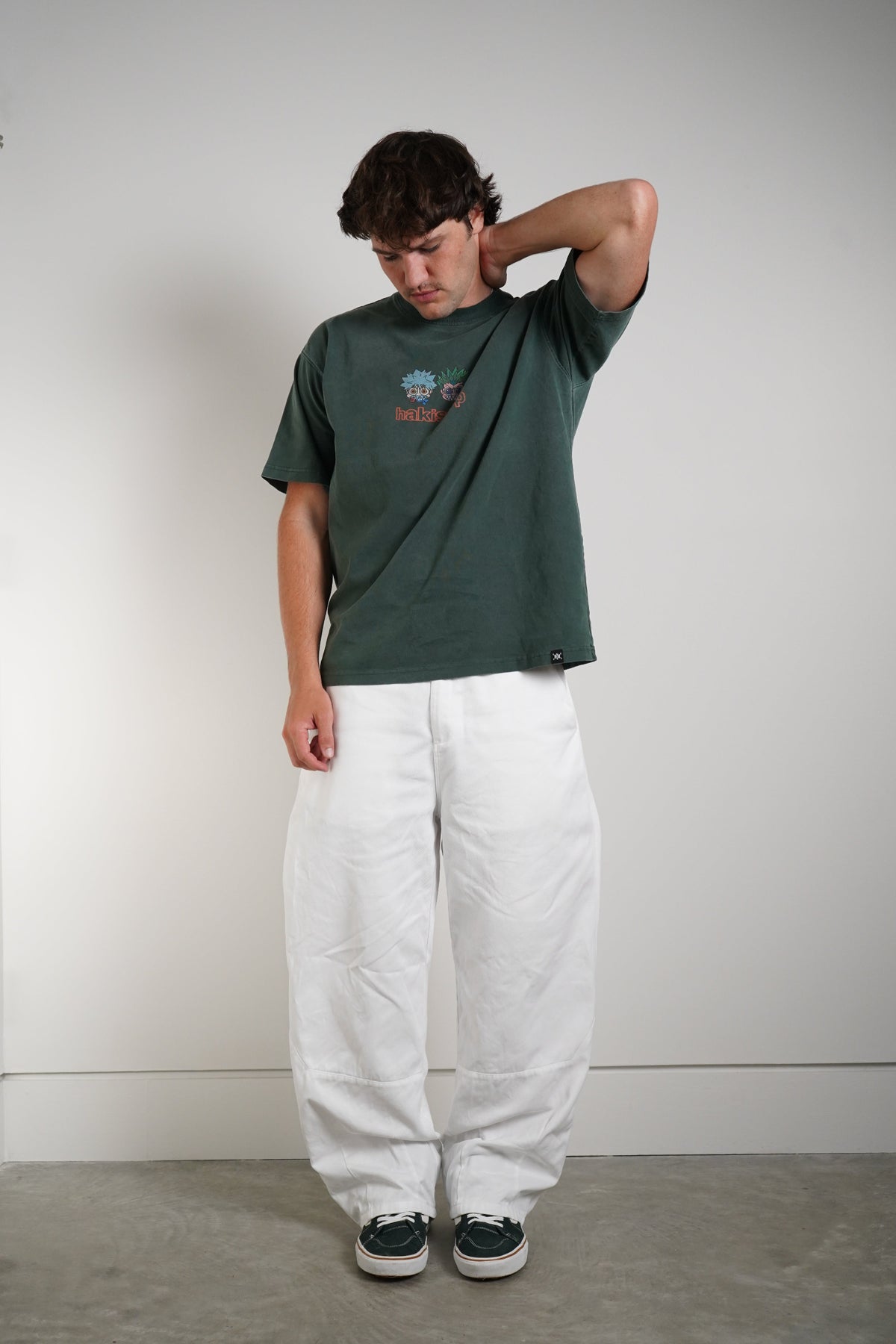 A man wearing a dark green graphic t-shirt, white baggy sweatpants, and black sneakers, standing against a plain light gray wall.