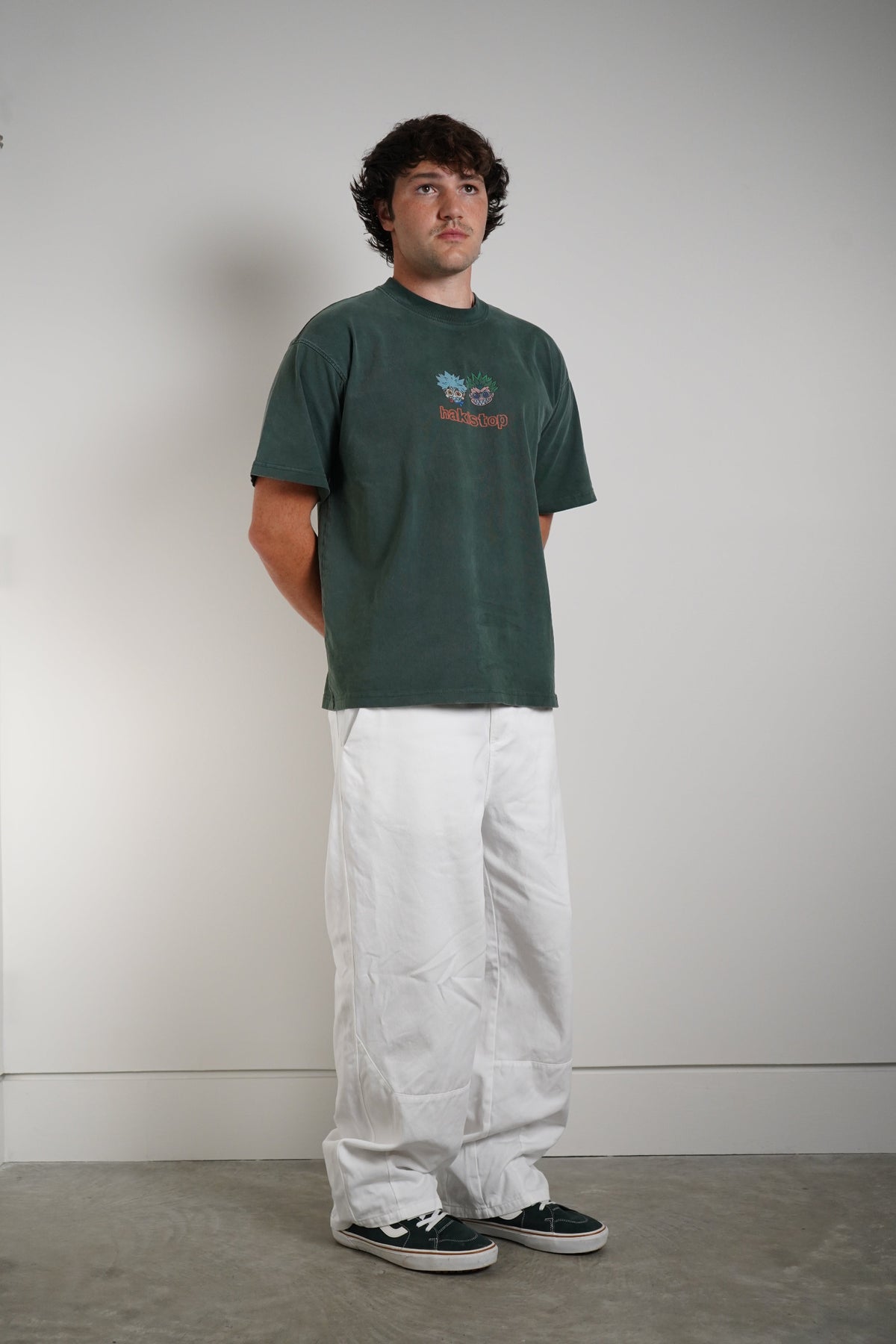 A man wearing a dark green graphic t-shirt, loose white pants, and dark green sneakers, standing against a plain light gray wall.