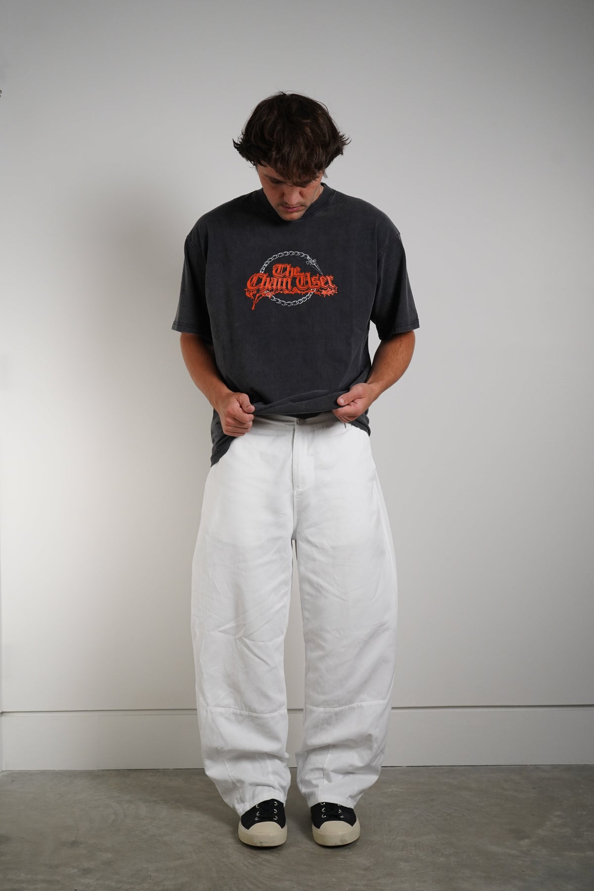 A person standing against a plain light gray wall wearing a faded black graphic t-shirt, loose white pants, and black-and-white sneakers.