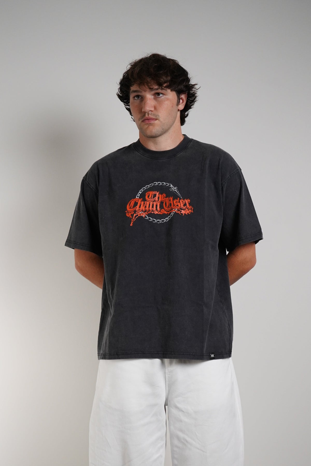 A man wearing a faded black graphic t-shirt with a orange and grey 'The Chain User' logo, paired with loose white pants and black-and-white sneakers, standing against a plain light gray wall.