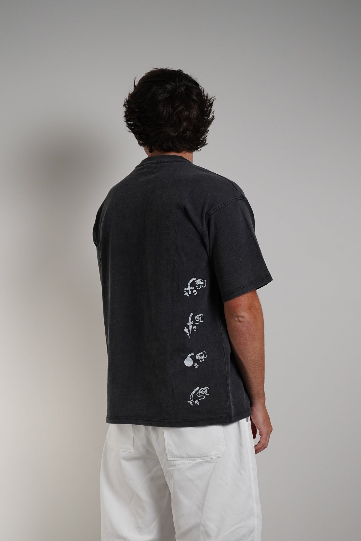 A man viewed from behind wearing a dark charcoal gray short-sleeve t-shirt with white prints down the back, paired with loose white pants and black sneakers.