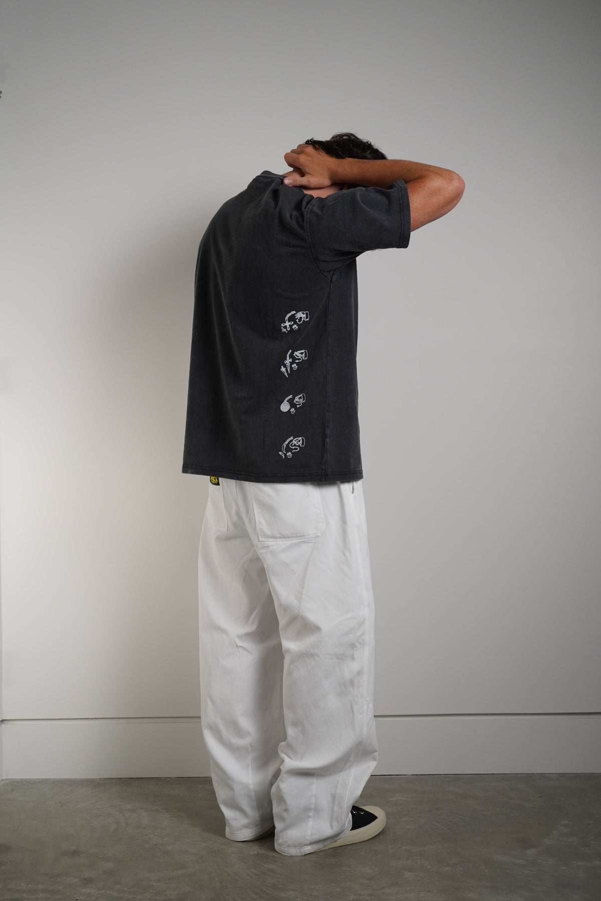 A person stands with their back to the camera, wearing a dark charcoal gray short-sleeve t-shirt with white embroidered details down the back and white wide-leg pants, set against a plain gray wall and concrete floor.
