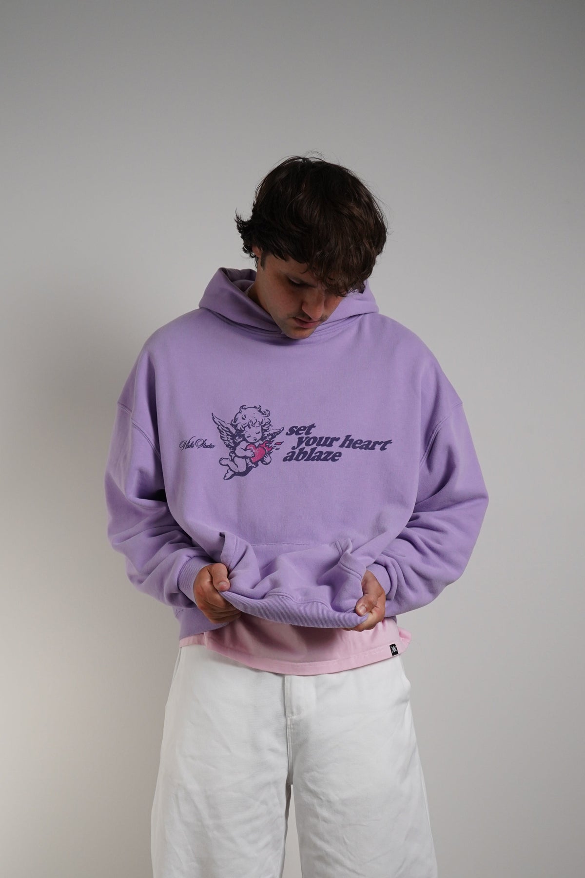 A person wearing a light purple hoodie with a graphic print on the chest, layered over a light pink shirt and paired with loose white pants and brown sandals, standing against a plain gray wall.
