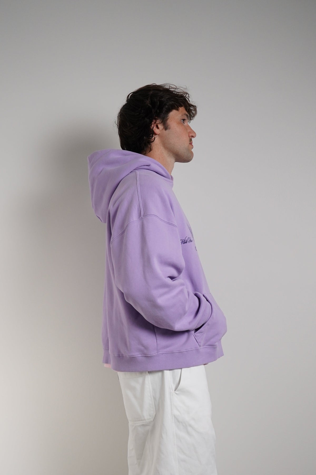 A man standing in profile wearing a lavender-purple oversized hoodie and loose white pants, photographed against a plain light gray wall and concrete floor.