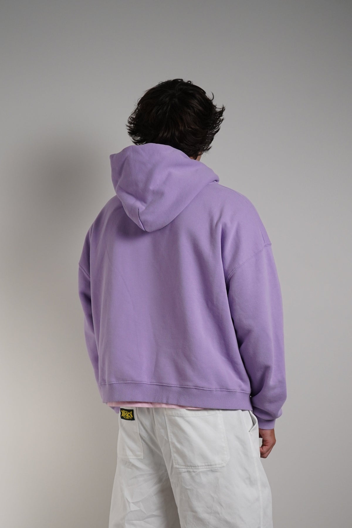 A person standing with their back to the camera, wearing a lavender purple hoodie and loose white pants, photographed in a minimalist studio setting with a gray wall and concrete floor.