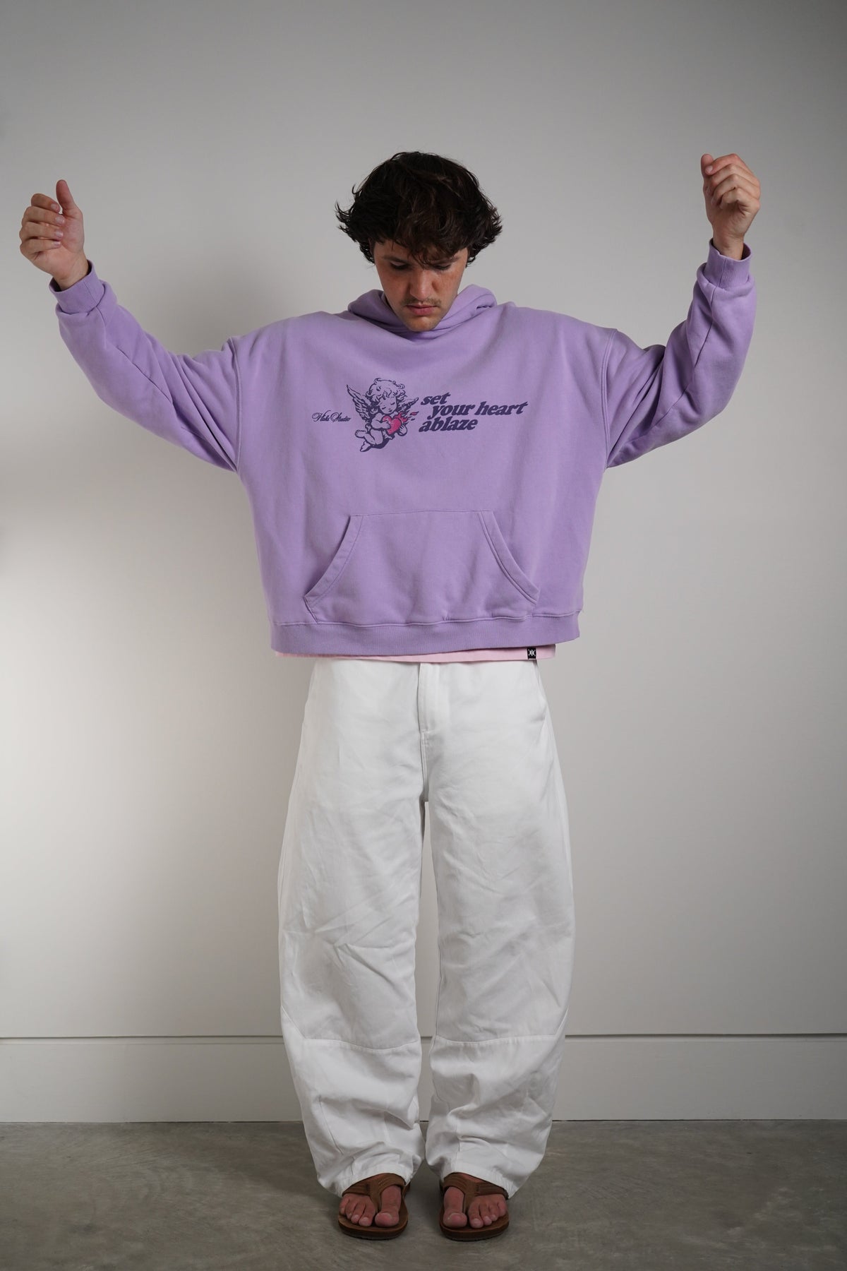 A person wearing a cropped lavender purple hoodie with a graphic print on the chest and loose white pants, standing against a plain light gray wall.