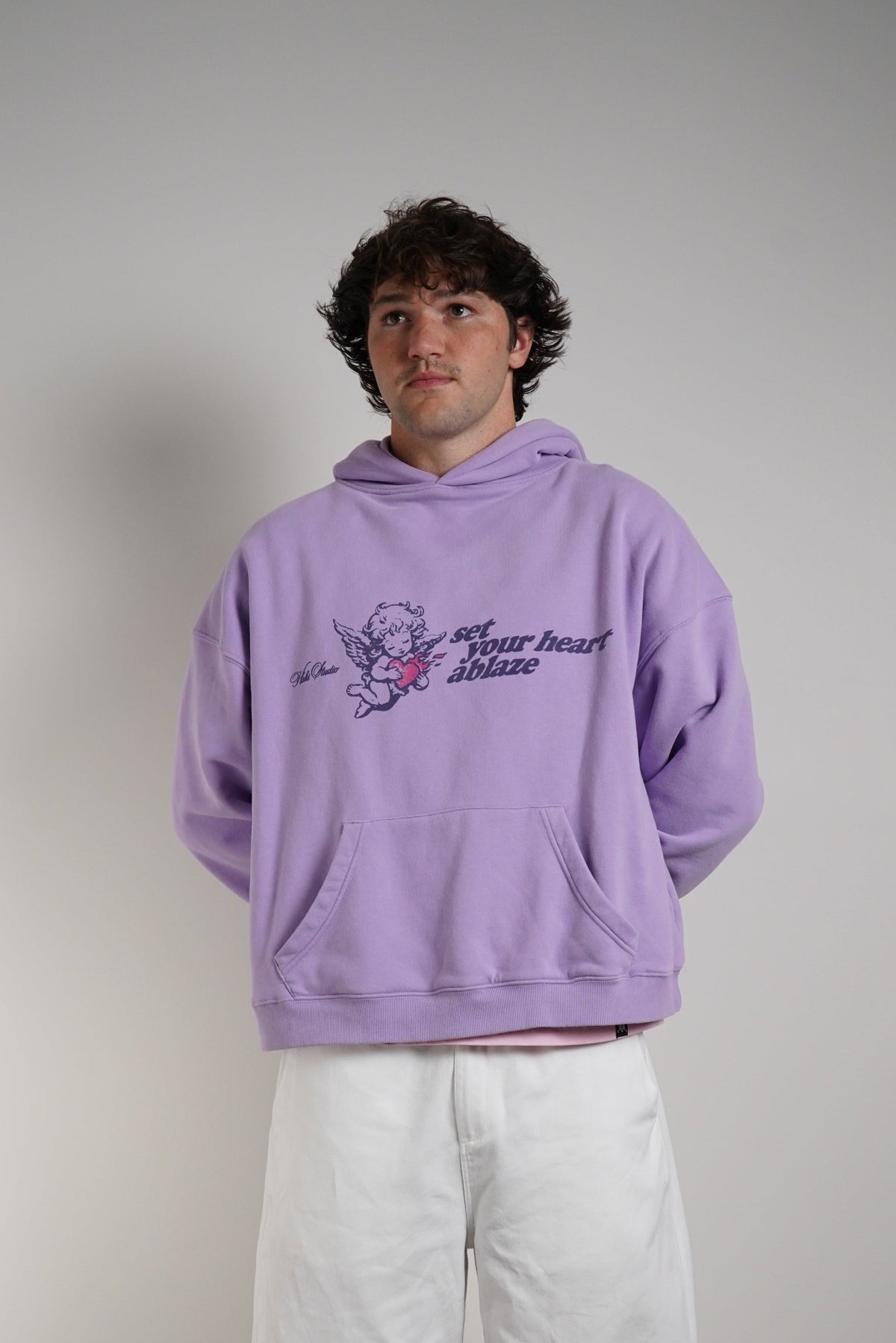 A man wearing a lavender purple hoodie with a graphic print on the chest and loose white pants, standing against a plain gray wall.