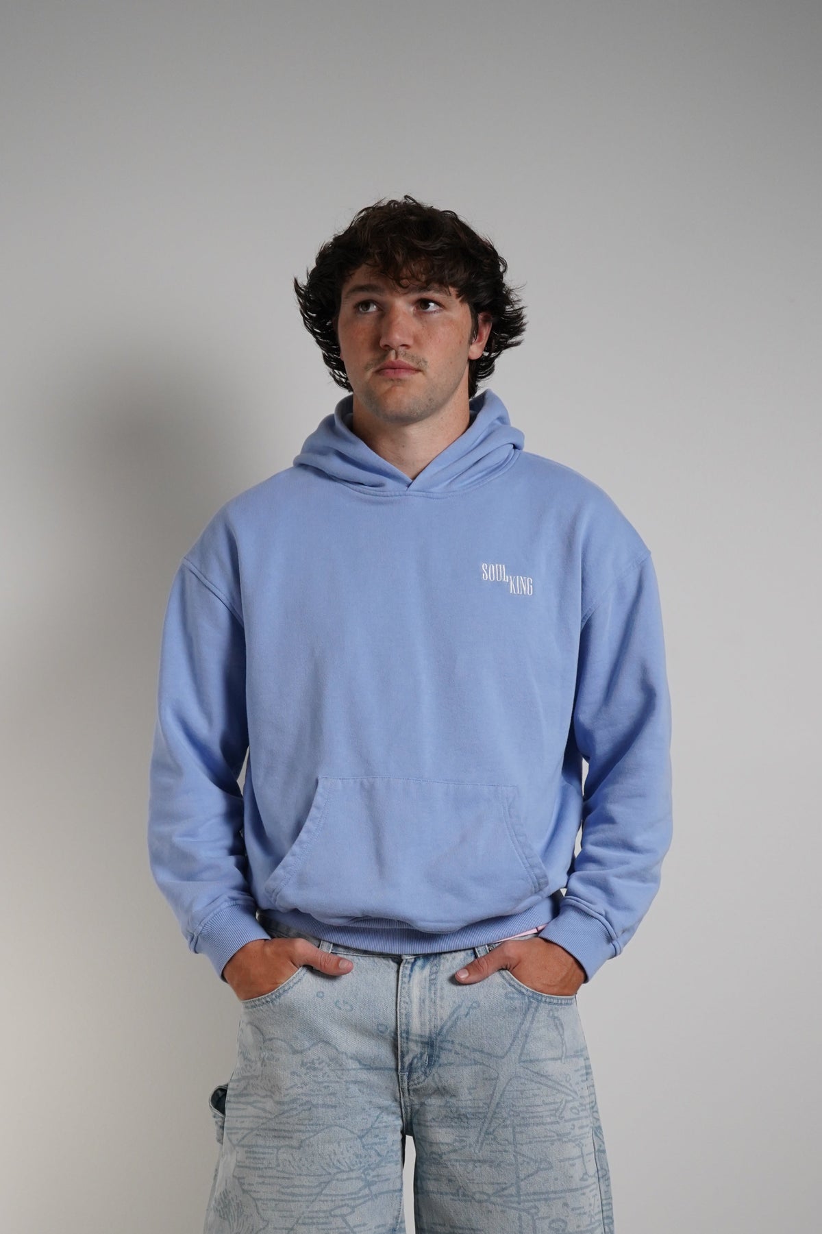 A man wearing a light blue hoodie with a small white chest logo and light-wash wide-leg jeans, standing against a plain light gray wall.