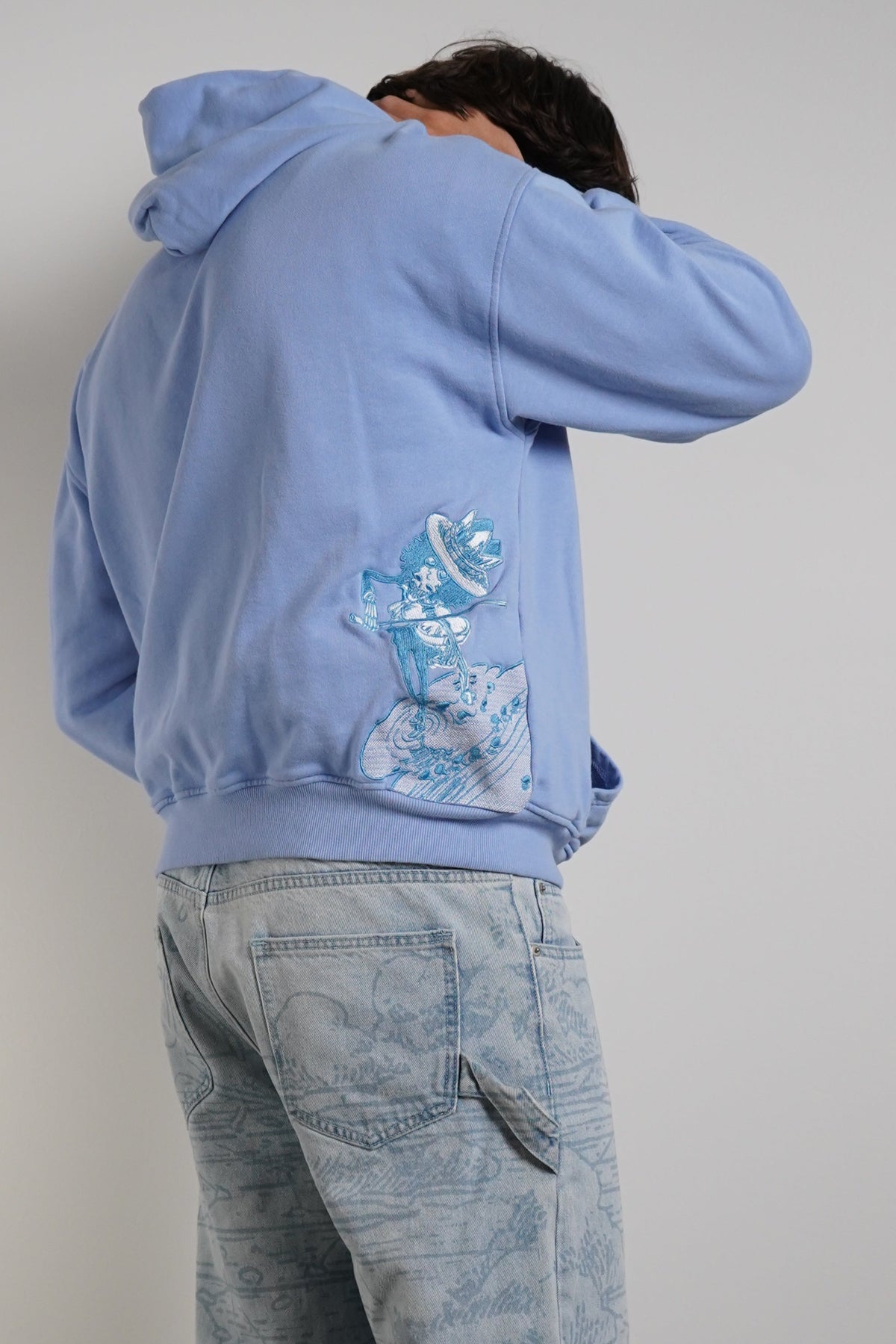 A person standing with their back to the camera, wearing a light blue hoodie with a blue graphic print on the back and light-wash denim jeans, set against a plain gray wall and concrete floor.