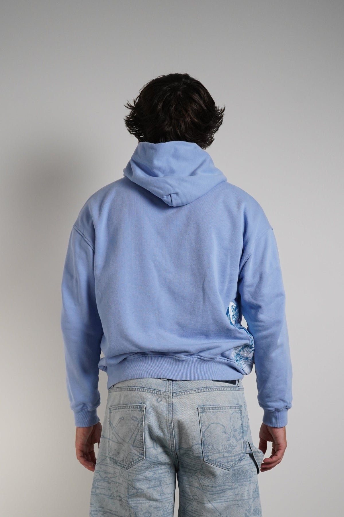 A person viewed from behind wearing a light blue hooded sweatshirt and very loose, wide-leg light blue jeans, standing against a plain light gray wall.