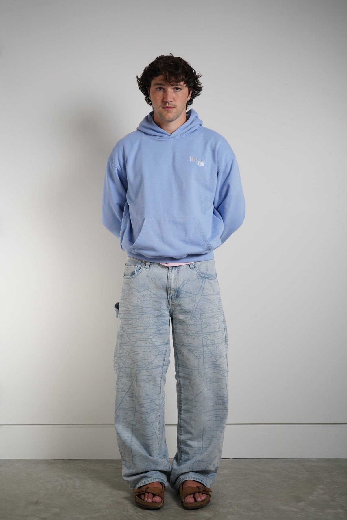 A man wearing a light blue hoodie with a small white logo on the left chest, paired with light-wash wide-leg jeans and brown sandals, standing against a plain light gray wall.