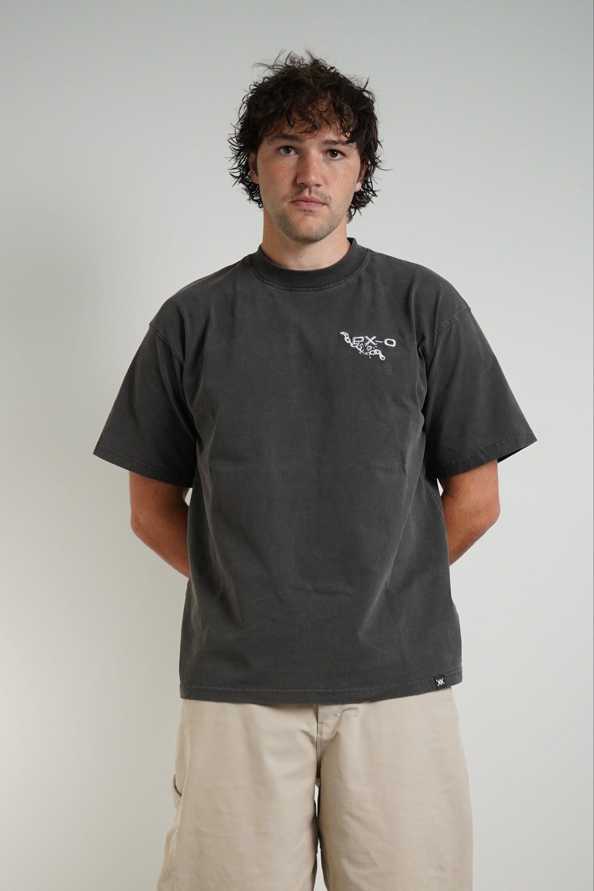 A man wearing a dark charcoal gray t-shirt with a small white logo on the left chest, paired with beige cargo pants and white sneakers, standing against a plain light gray wall.