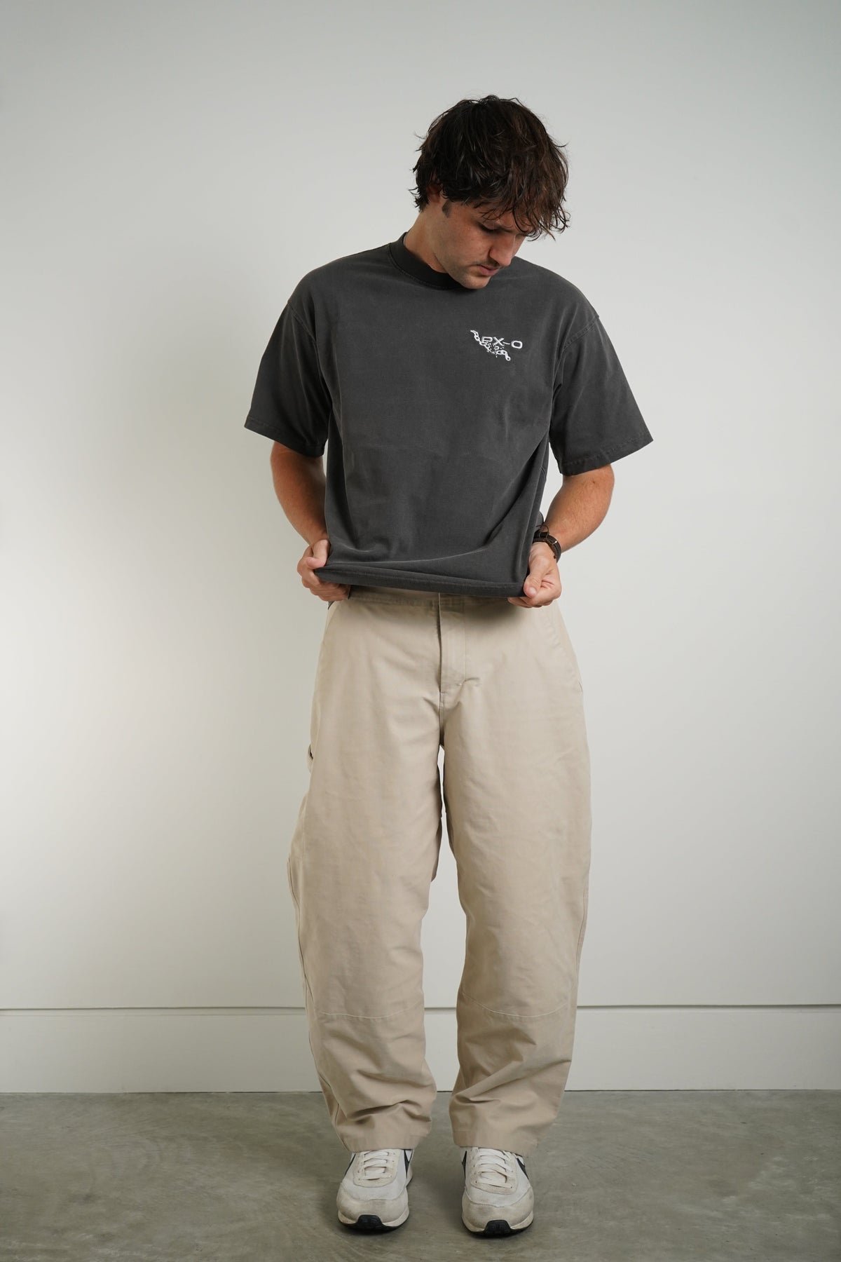 A man wearing a dark gray oversized t-shirt with a small white graphic on the left chest, paired with beige cargo pants and white sneakers, standing against a plain light gray wall.