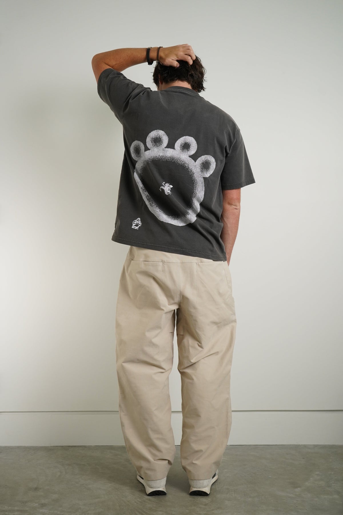 A man viewed from behind wearing a dark gray t-shirt with a large white paw print graphic on the back, paired with beige cargo pants and white sneakers, standing against a plain light gray wall.