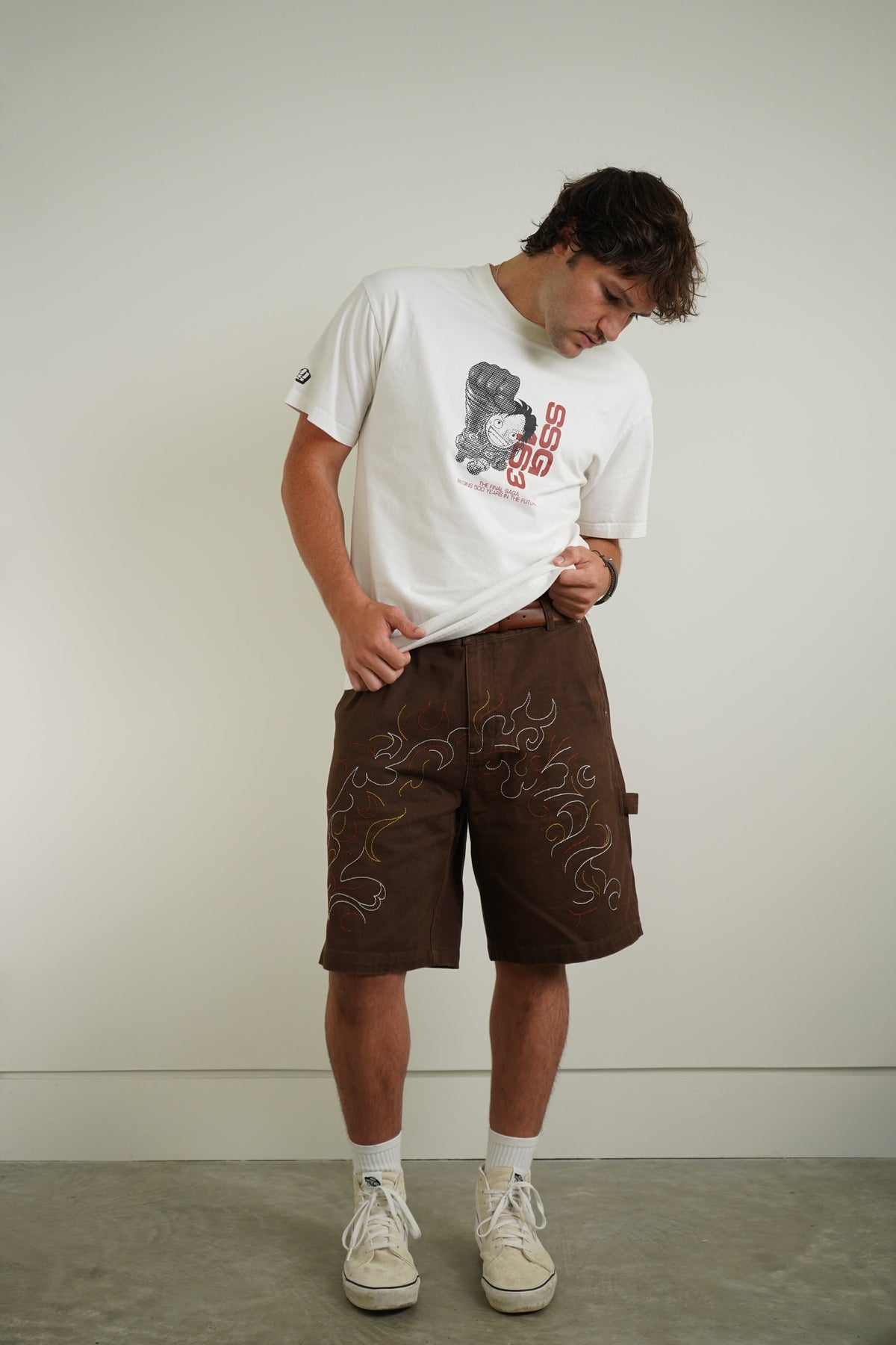 A man wearing a white graphic t-shirt, brown shorts with embroidered details, white socks, and off-white sneakers, standing against a plain light gray wall.