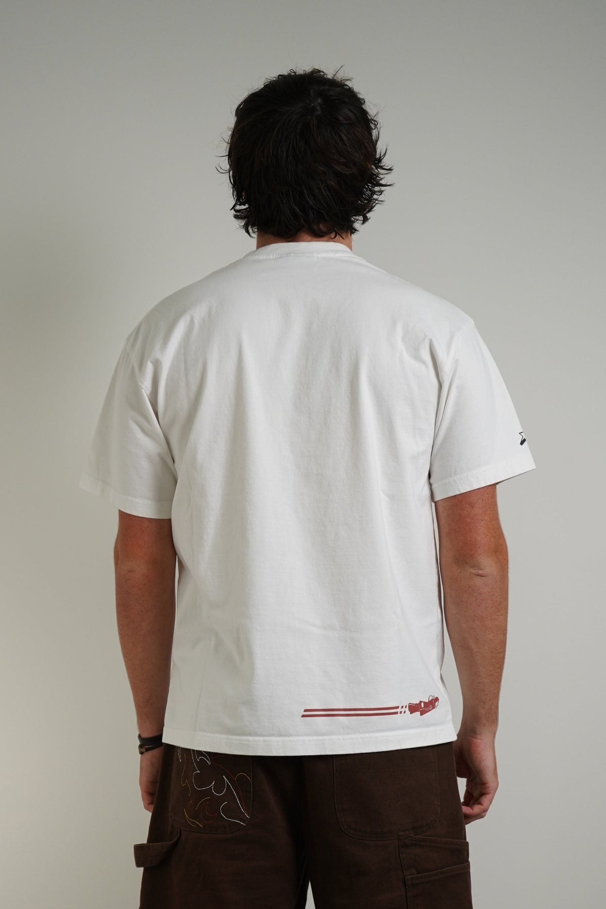 A man viewed from behind wearing a white t-shirt with a small red graphic on the lower back, dark brown cargo shorts, white socks, and white sneakers, standing against a plain light gray wall.