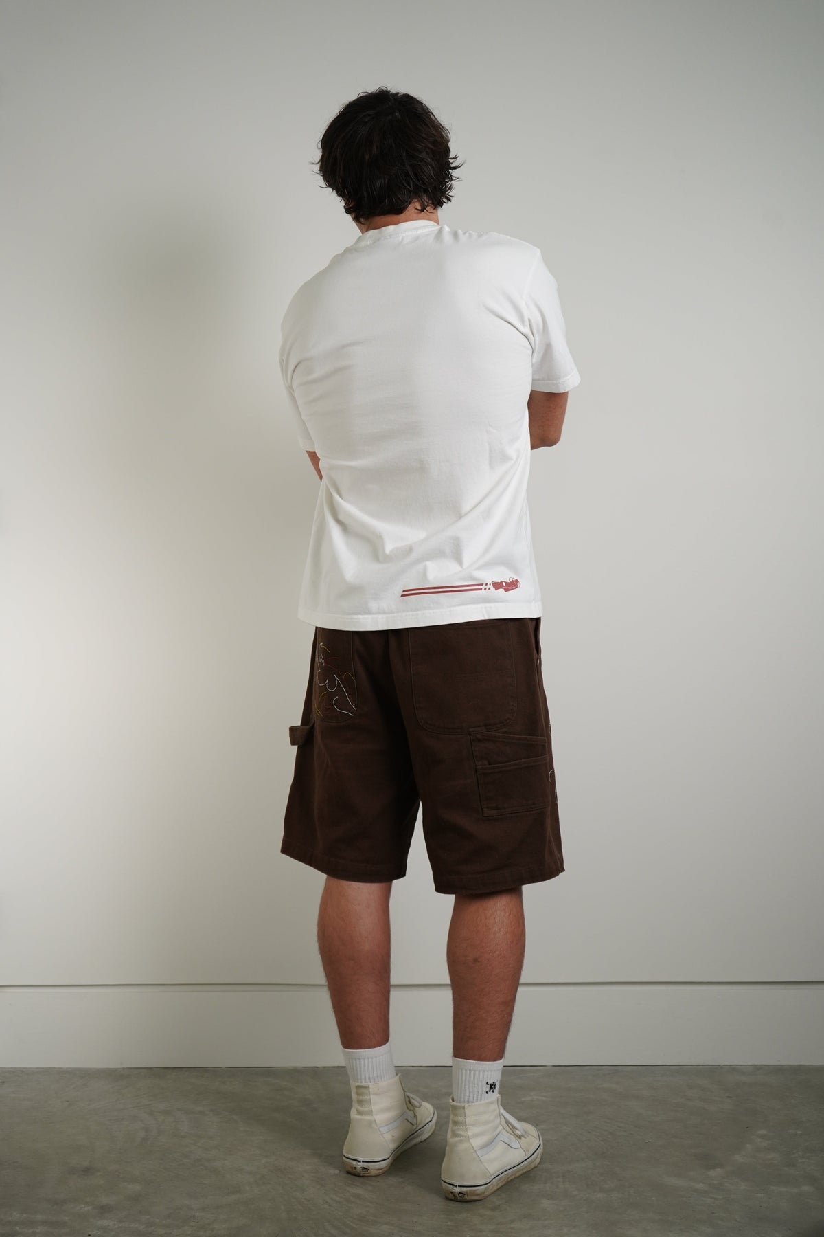 A man viewed from behind wearing a white t-shirt with a small red graphic on the lower back, dark brown cargo shorts, white socks, and white sneakers, standing against a plain light gray wall.