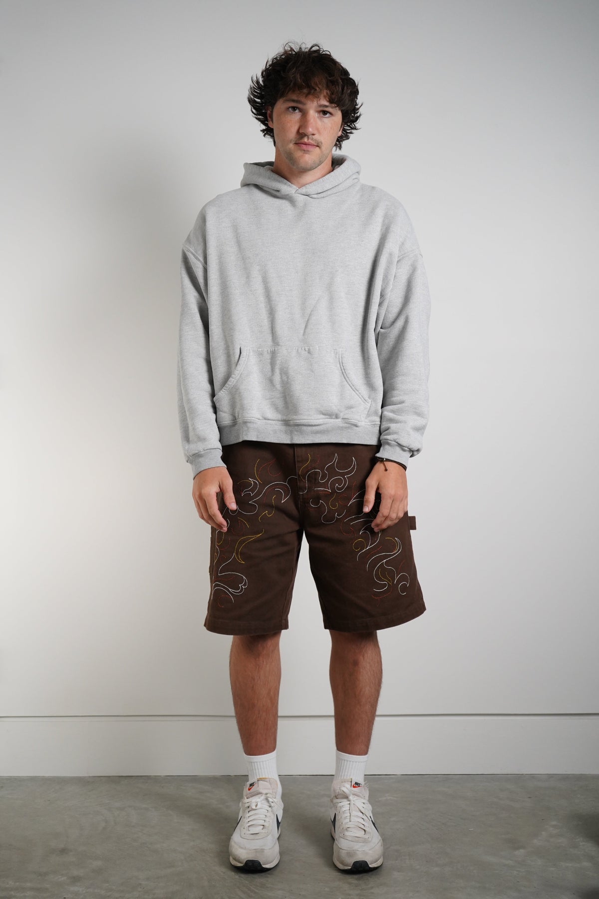 A man stands against a plain light gray wall wearing a light gray pullover hoodie, brown shorts with white embroidered swirl patterns, white socks, and white sneakers.