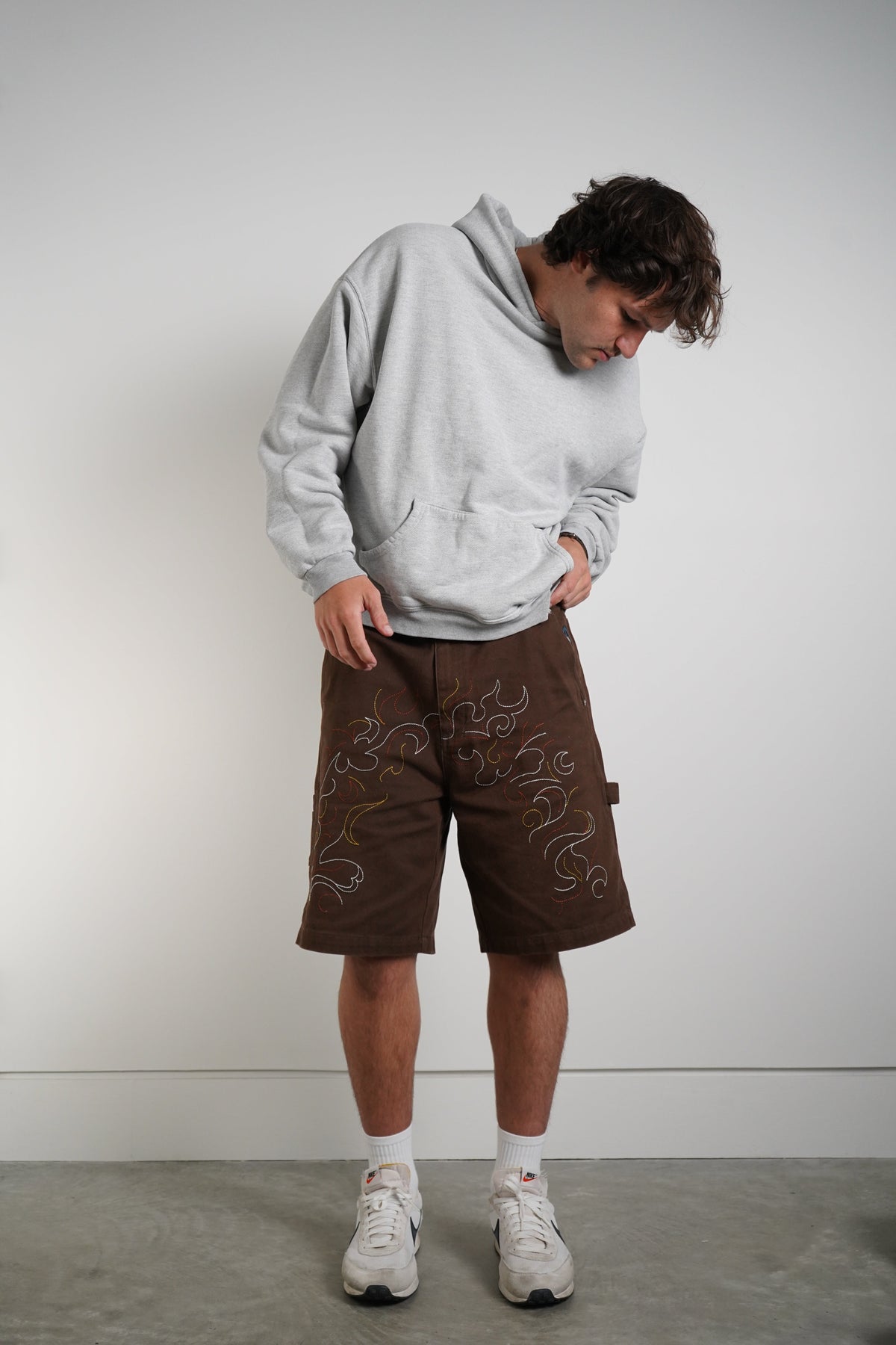 A man wearing a light gray hoodie, brown shorts with white embroidered swirl patterns, white socks, and white sneakers, standing against a plain light gray wall.