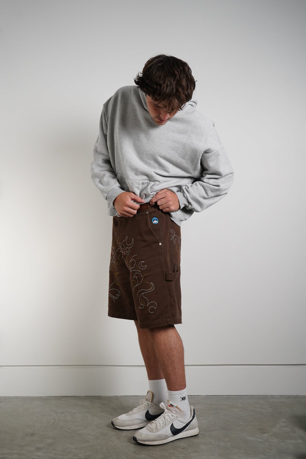 A person wearing a light gray sweatshirt, brown embroidered shorts, white socks, and white Nike sneakers, standing against a plain light gray wall.