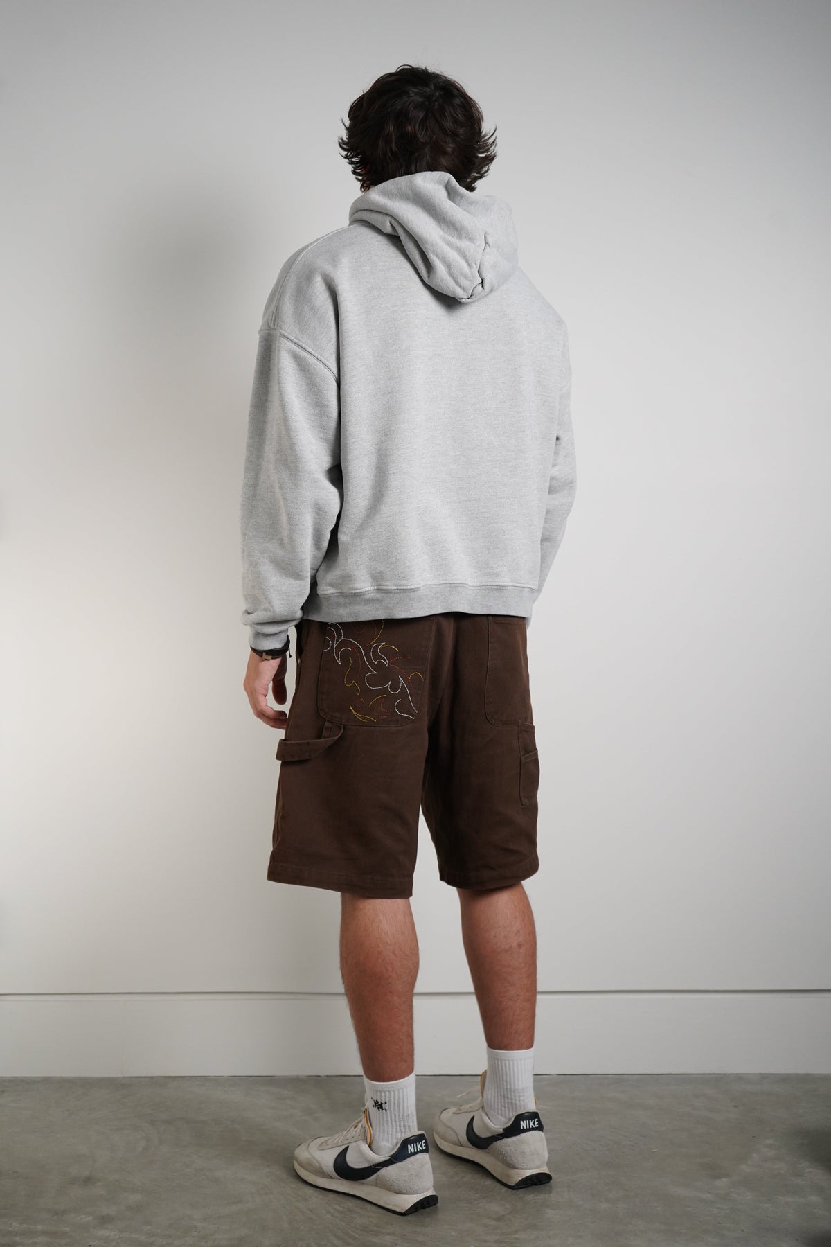 A man is shown from behind wearing a light gray heathered hoodie, dark brown cargo shorts with embroidery, white socks, and white sneakers with dark blue accents, standing against a plain light gray wall.