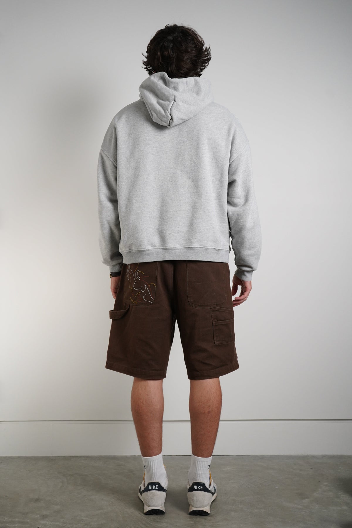 A man is shown from behind wearing a light gray heathered hoodie, brown cargo shorts with a graphic print, white socks, and sneakers, standing against a plain light gray wall.