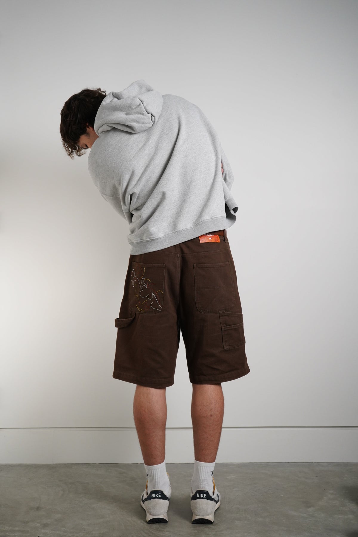 A person wearing a light gray oversized hoodie and brown cargo shorts, viewed from behind against a plain light gray wall and concrete floor.