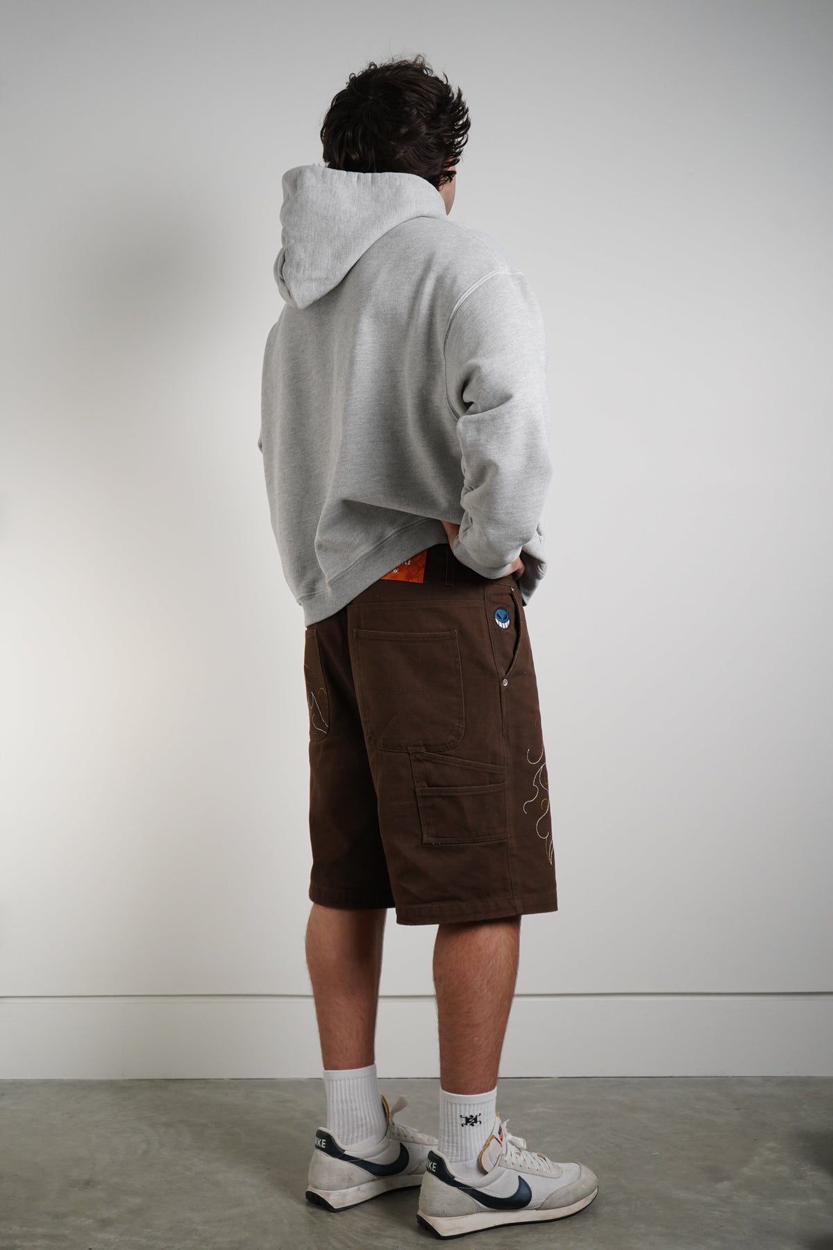 A person shown from the back wearing a light gray oversized hoodie, brown cargo shorts, white socks, and white sneakers with blue accents, standing against a plain light gray wall.