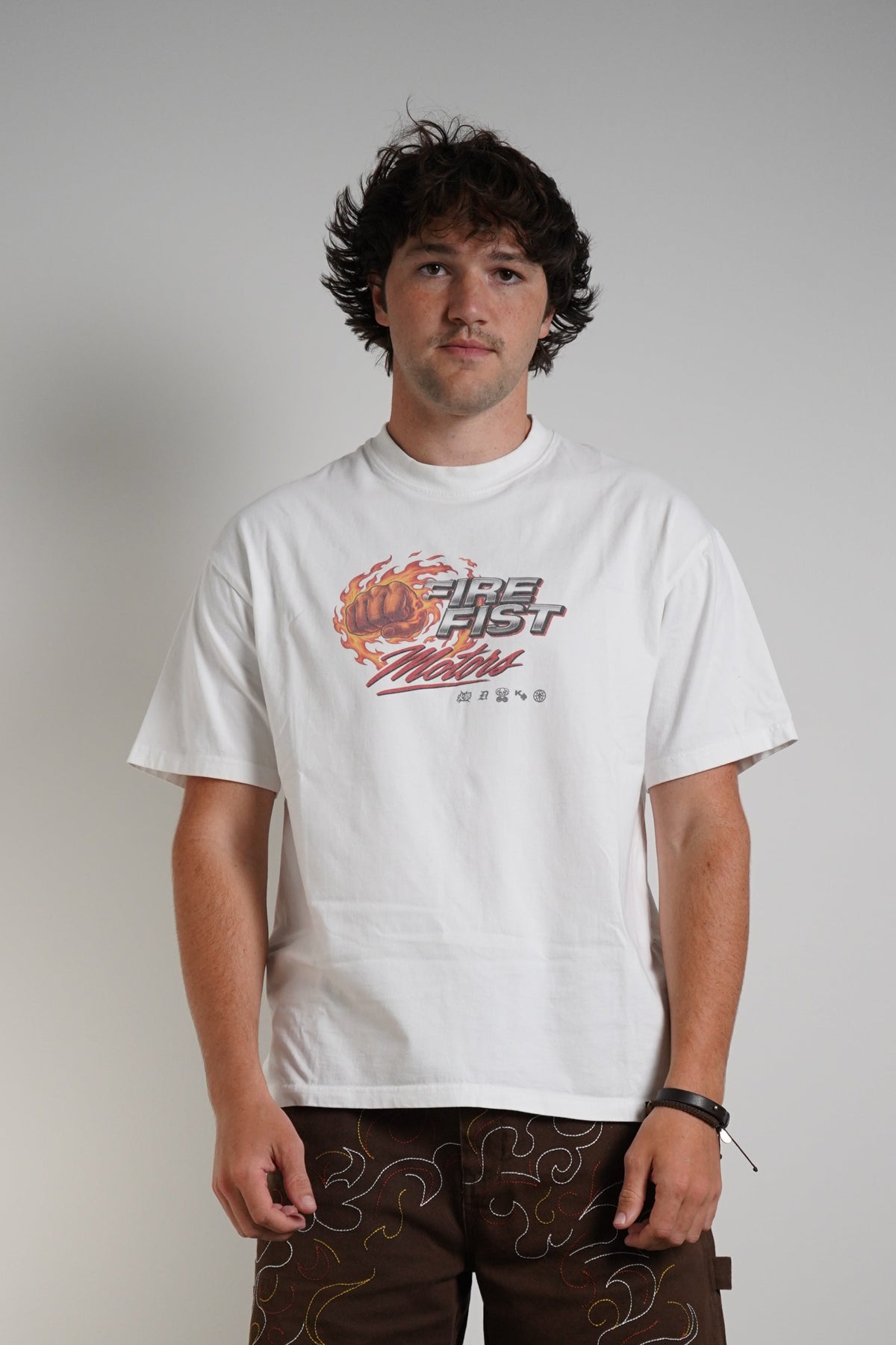 A man stands against a plain light gray wall wearing a white graphic T-shirt, dark brown shorts with a subtle pattern, white socks, and white sneakers.