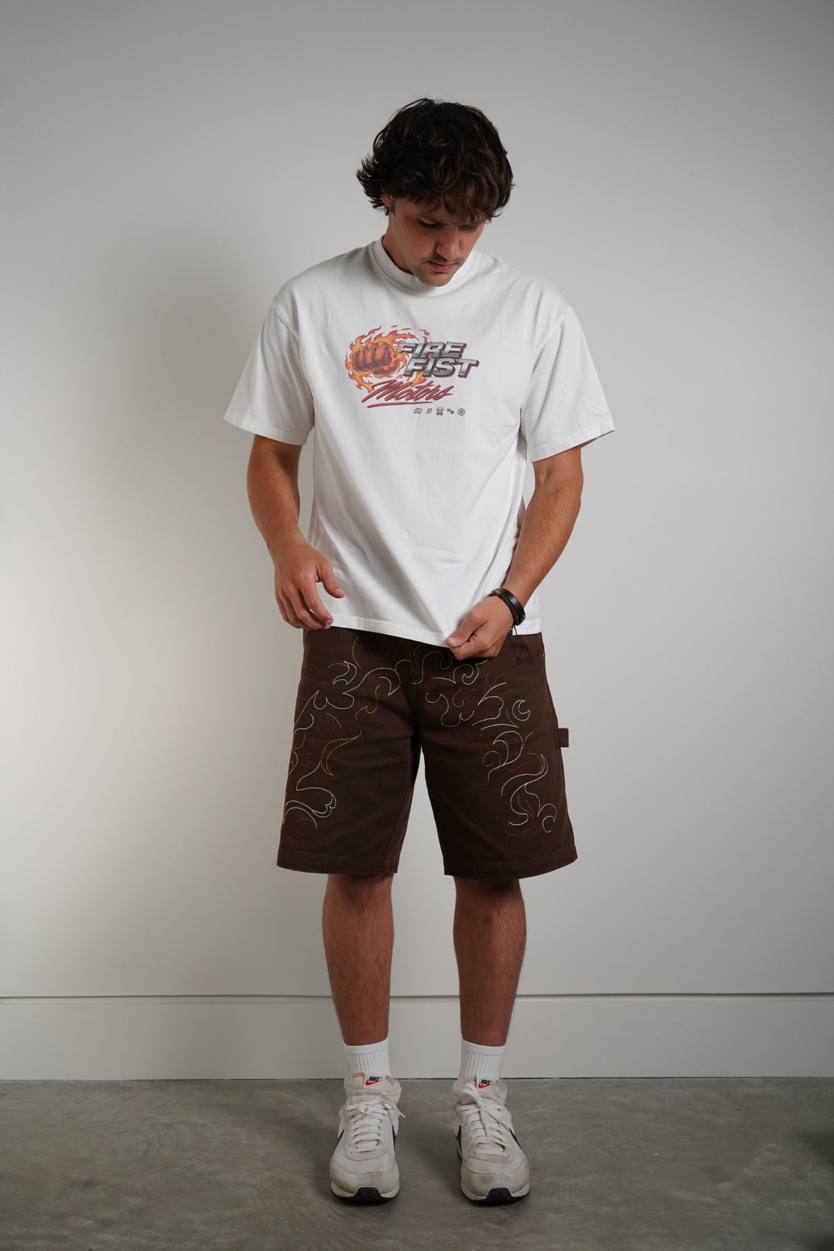 A man modeling a white graphic t-shirt and brown shorts with white socks and sneakers, standing against a plain gray studio wall.